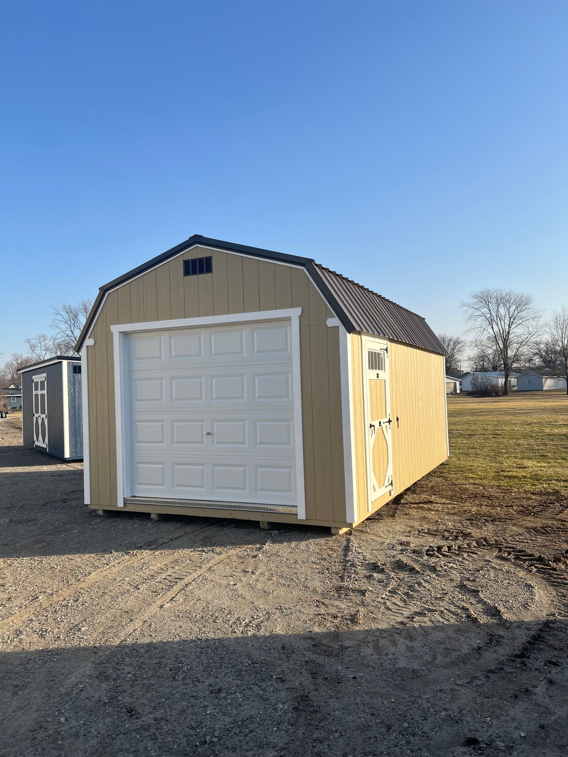 A tan storage shed with a white garage door, a side pedestrian door, and a brown metal gambrel roof on a gravel lot.