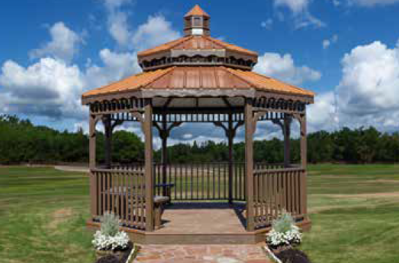 Wooden gazebo with copper-colored roof, on a brick pathway, with flower pots, set in a grassy field under blue sky.