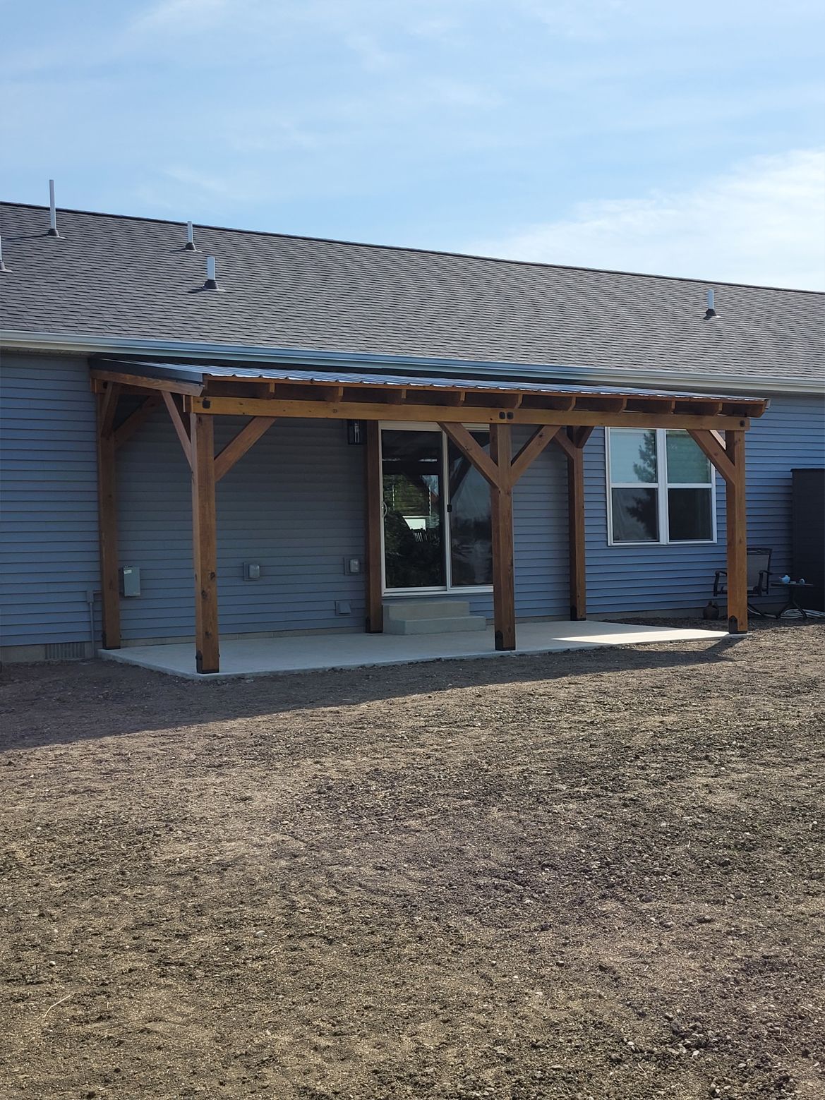 A wooden pergola attached to the rear of a blue-sided house with a concrete patio and a gravel yard.