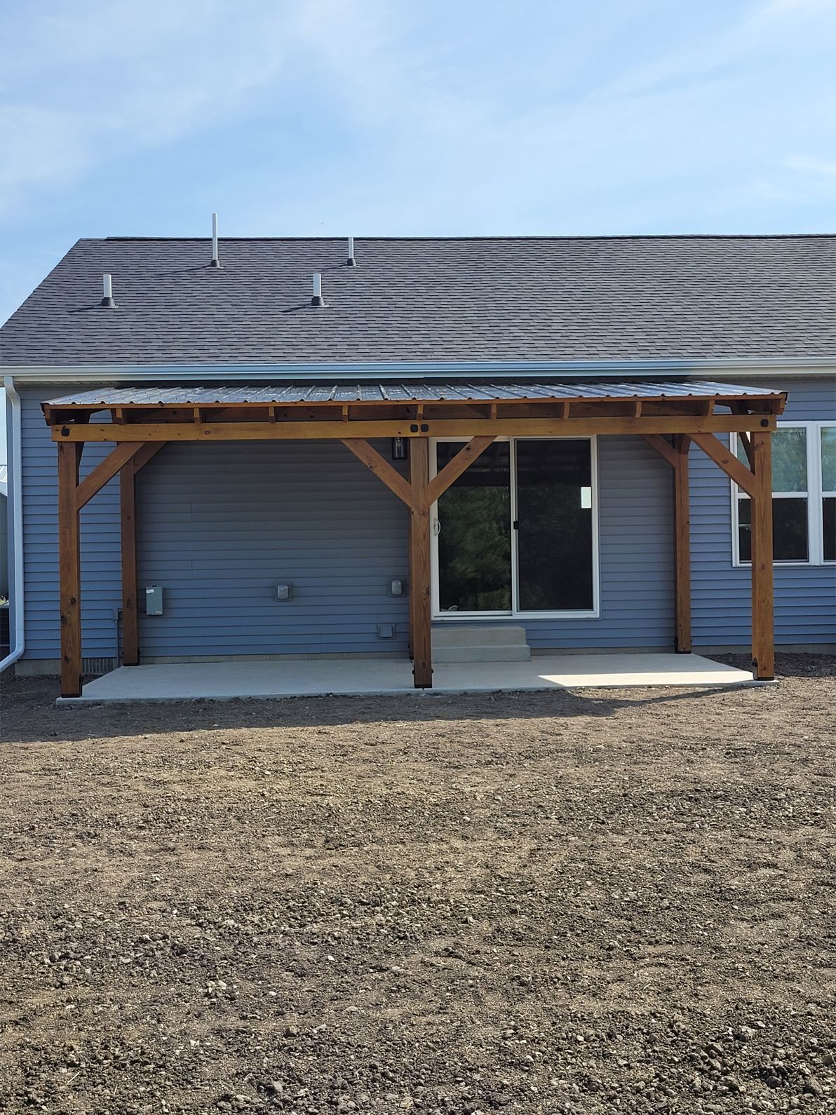 A wood-framed patio cover attached to the back of a blue-sided house with a gravel yard in the foreground.