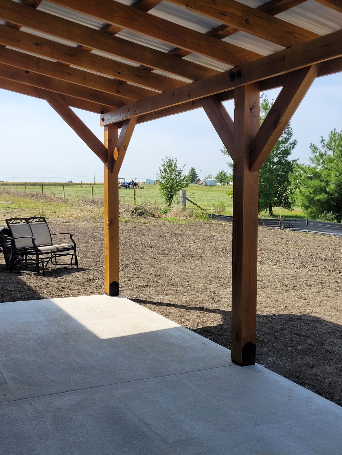 A covered concrete patio with wooden posts and a view of a grassy field with trees and a vehicle in the distance.