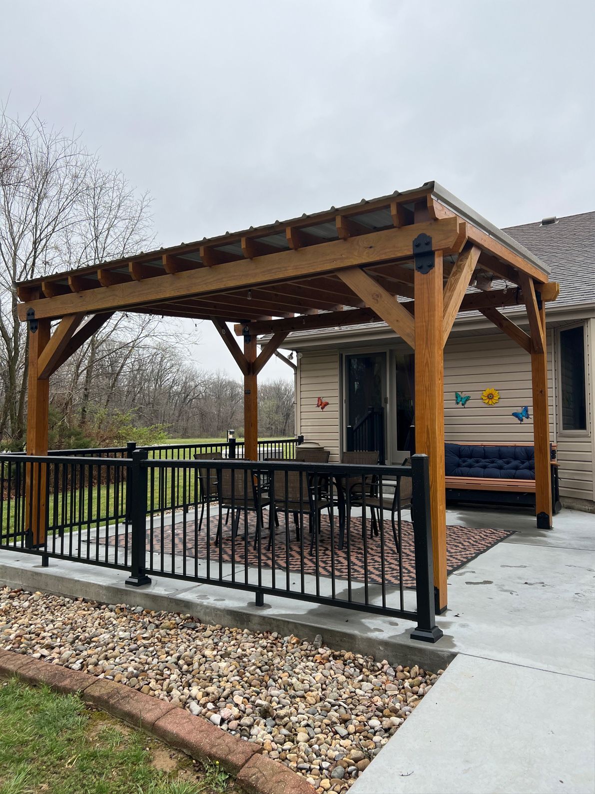 A wooden pergola with a dark metal roof stands over an outdoor patio set on a concrete slab near a house.