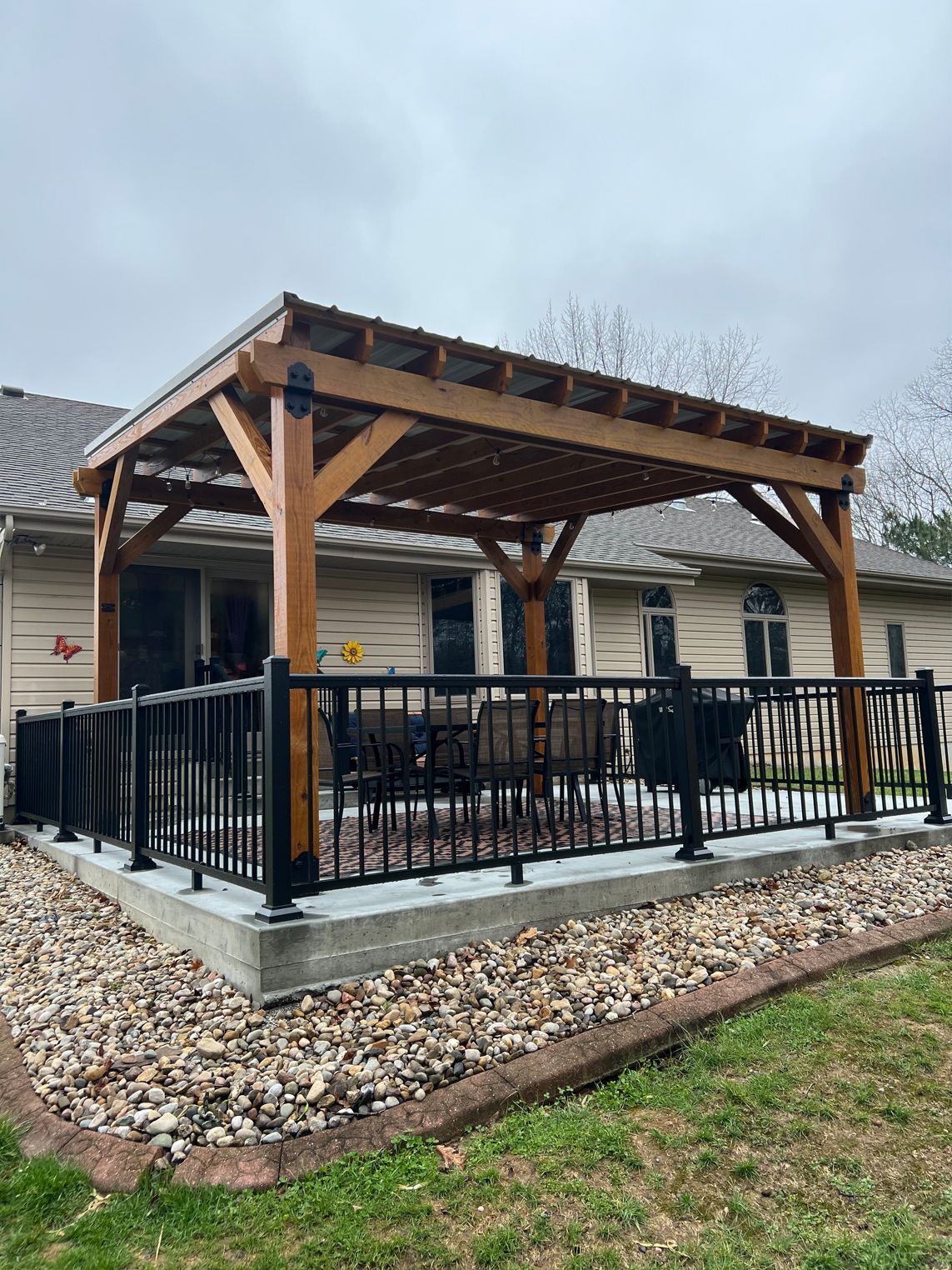 A wooden pergola with a dark metal roof stands on a concrete patio, surrounded by a black metal railing and gravel.