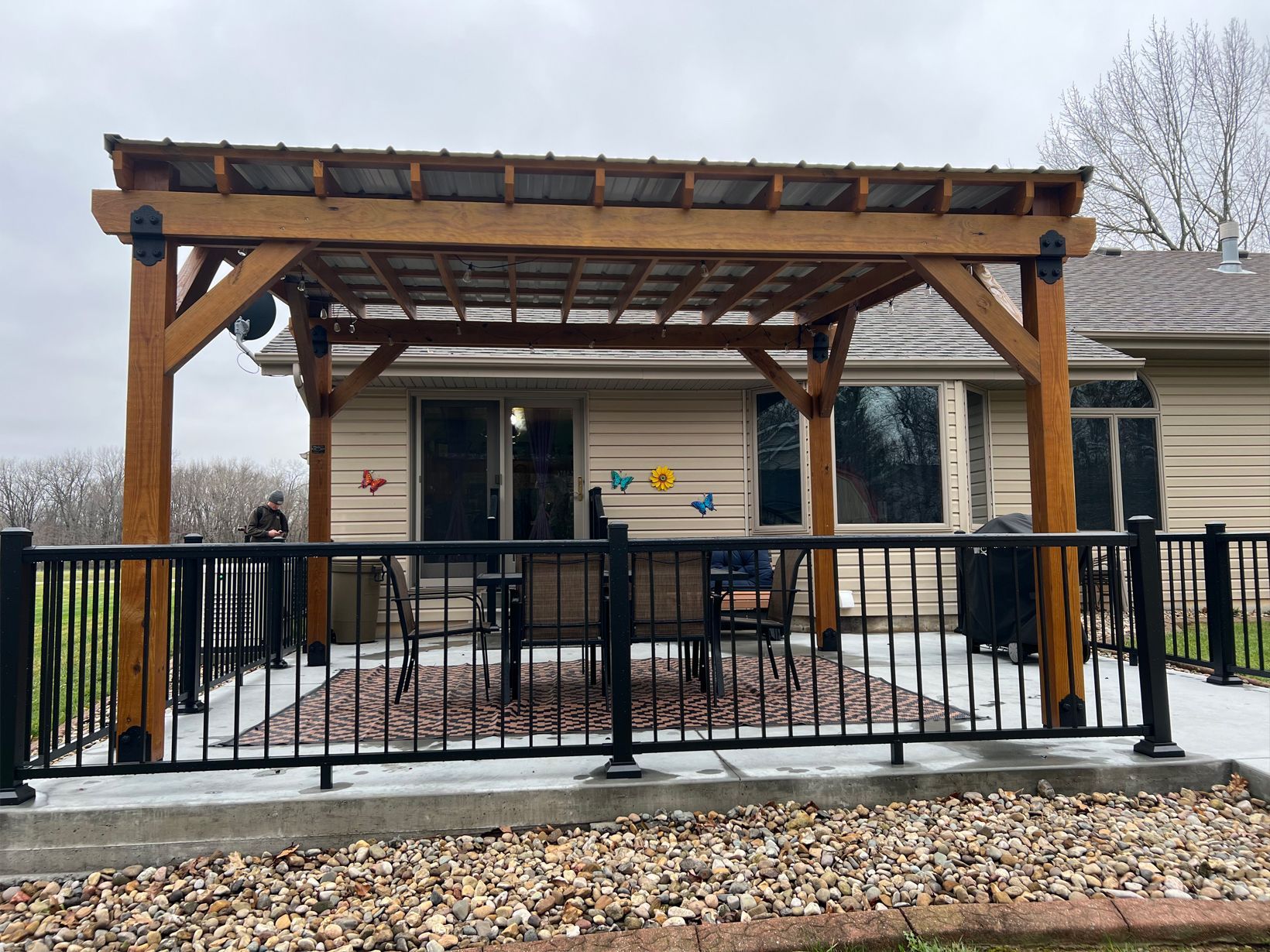 A wooden pergola with a metal roof covers a concrete patio with a dining set, surrounded by a black metal railing.