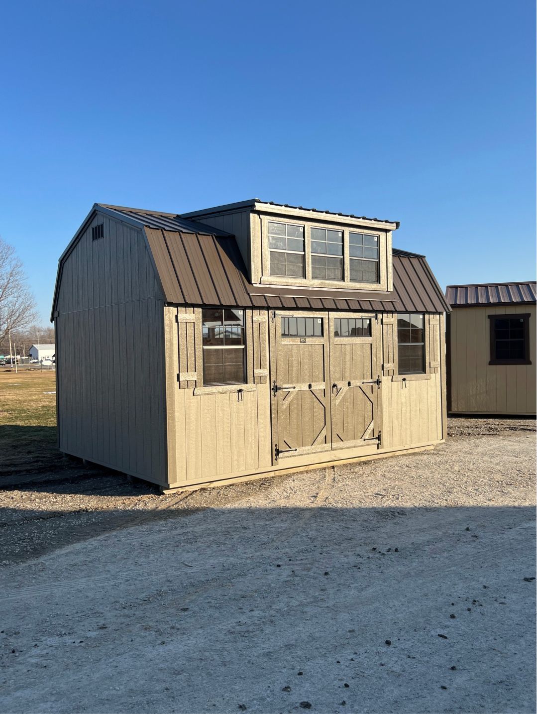 A beige wooden storage shed with a brown metal roof, double doors, and a dormer window, sitting on a gravel lot.