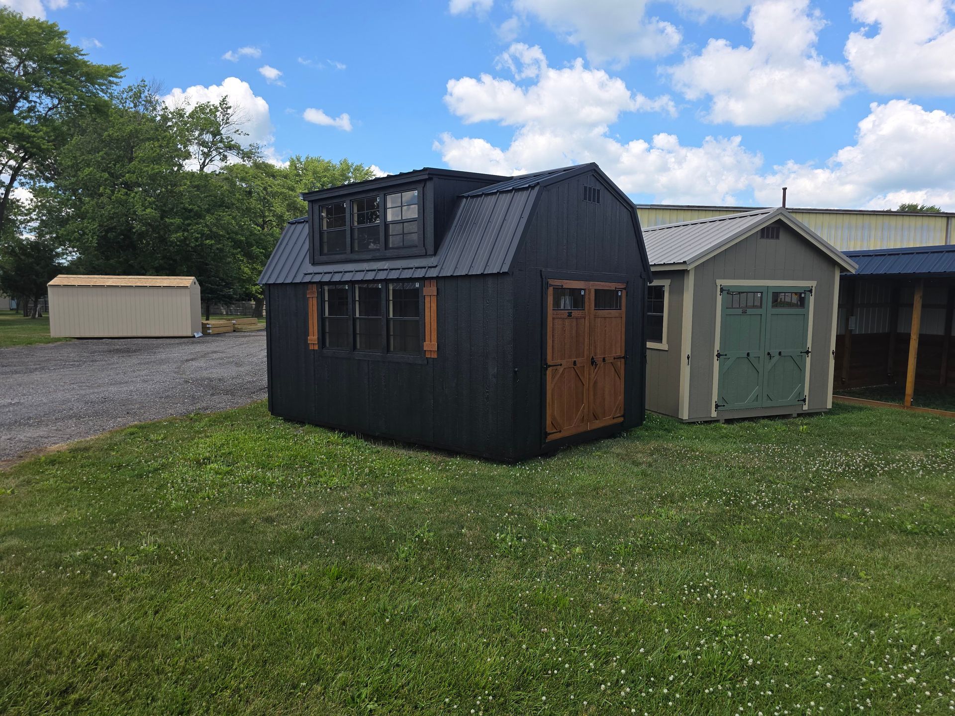 A black shed with a dormer window sits on a grassy field next to a beige shed under a partly cloudy sky.
