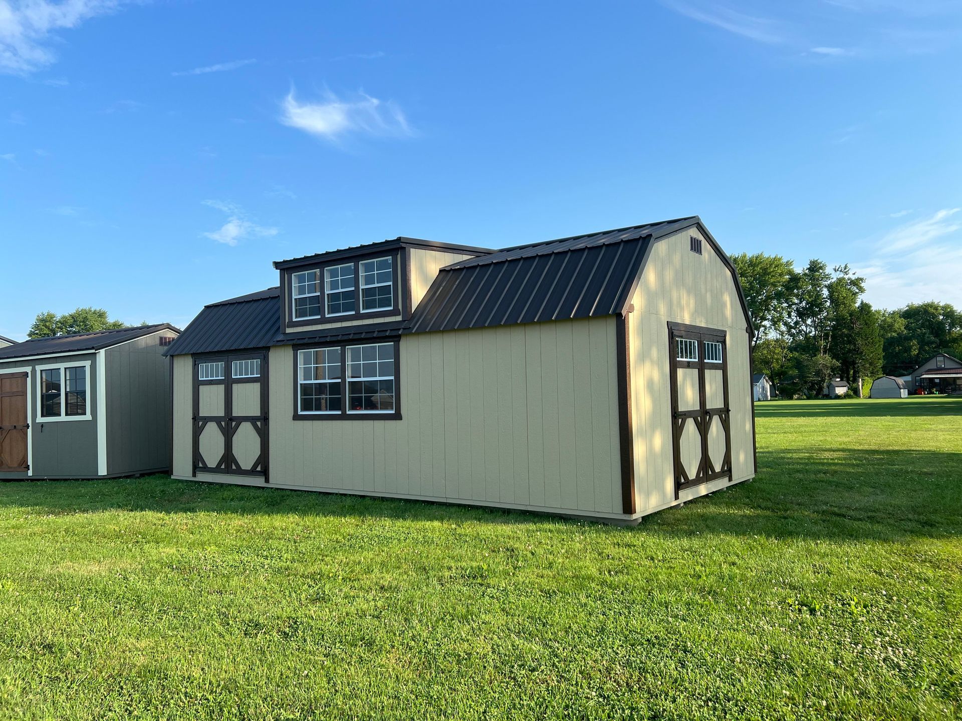 A tan wooden shed with a dark metal roof, dormer windows, and double doors, standing in a sunny, grassy field.