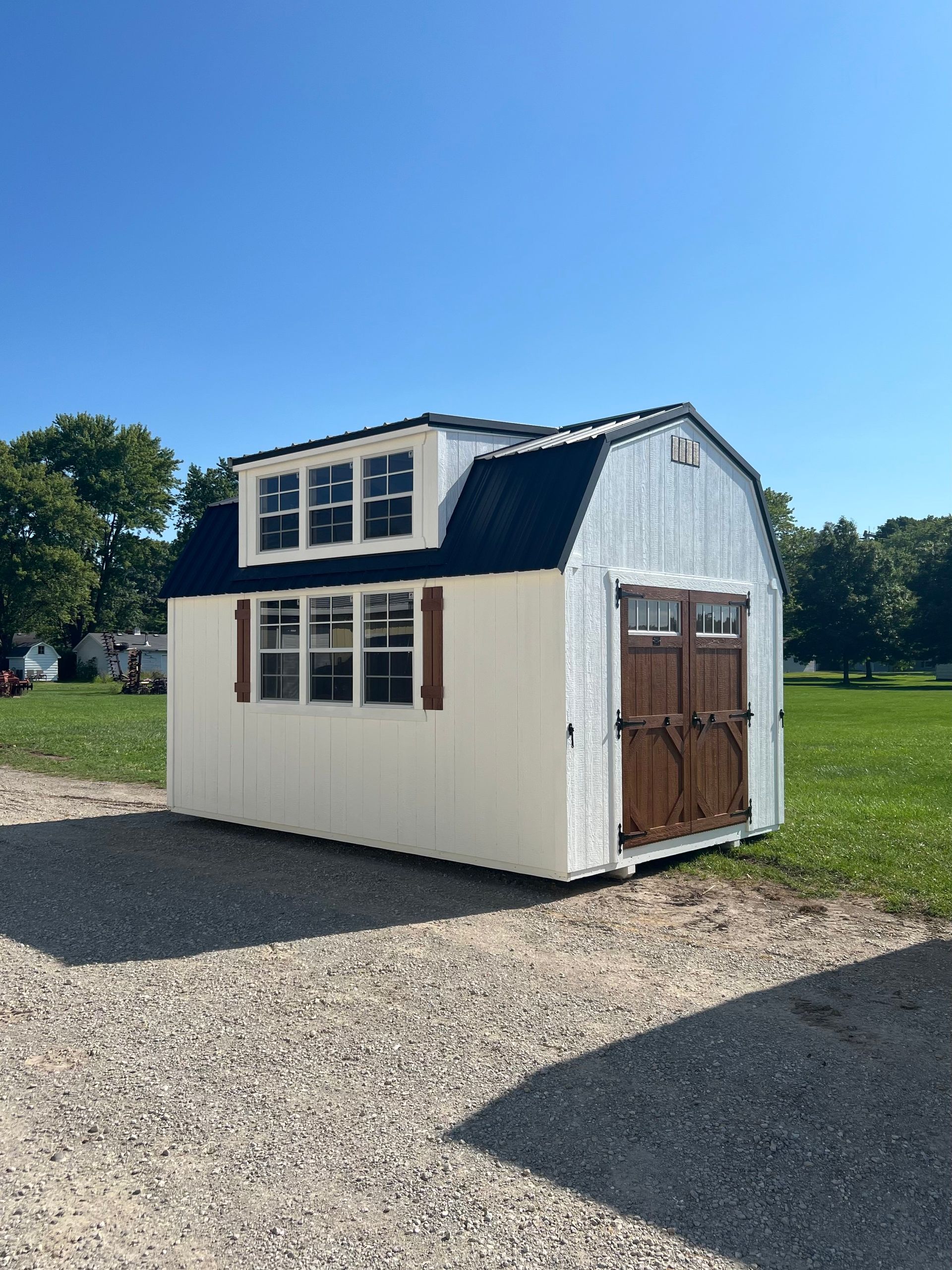 A cream-colored shed with a black gambrel roof, dormer windows, brown shutters, and double doors on a gravel lot.