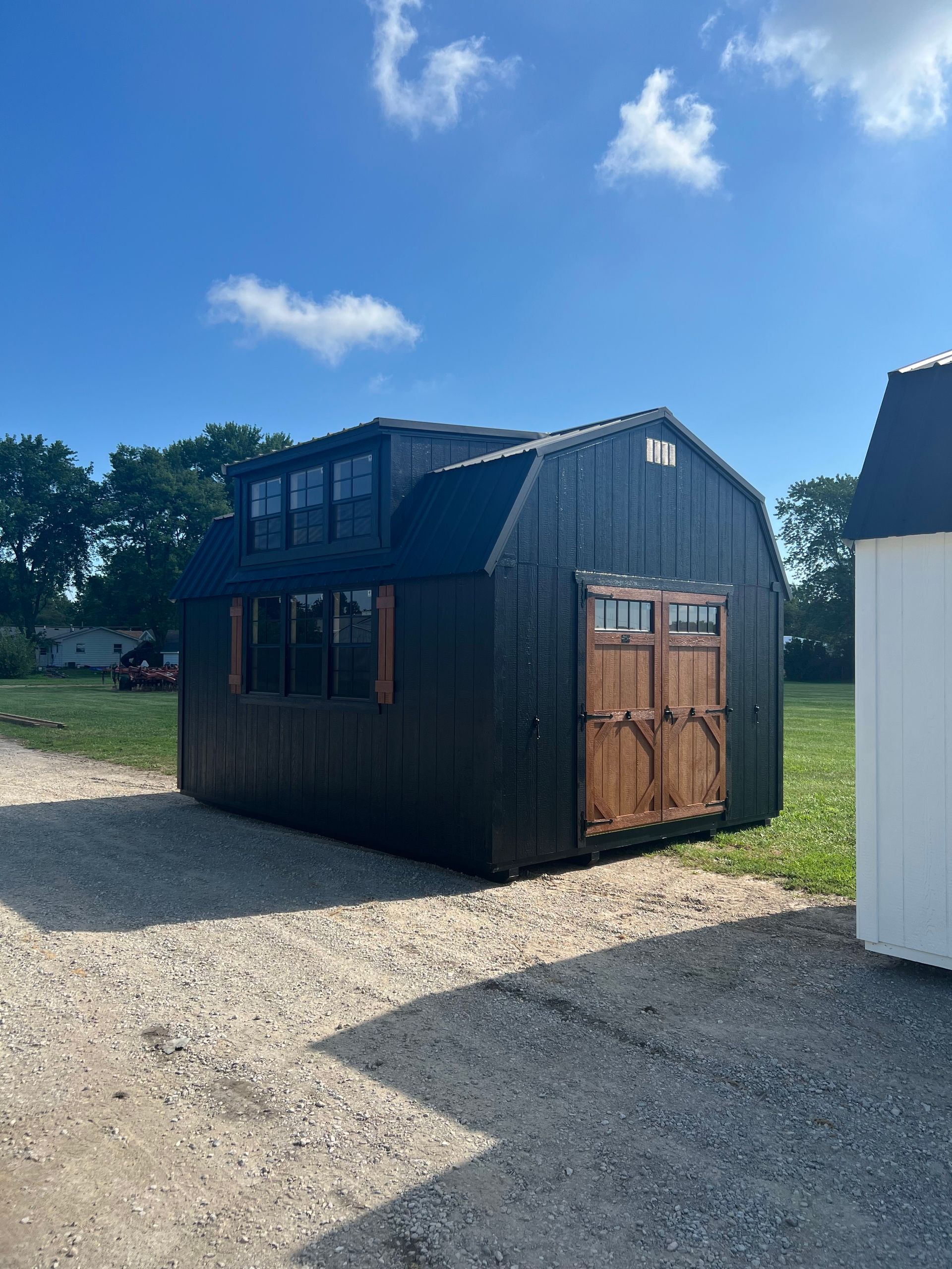 A black, barn-style shed with wooden double doors and a loft window sits on a gravel lot under a bright blue sky.