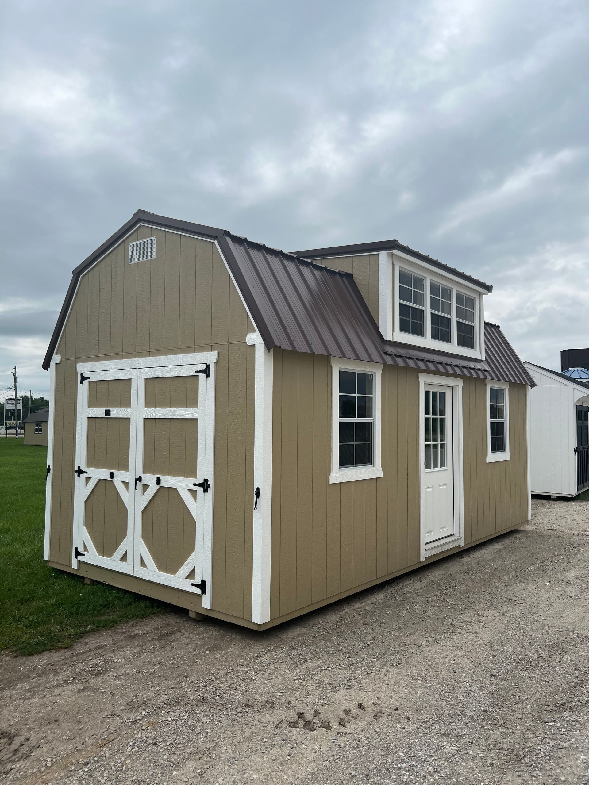 A tan barn-style shed with a dark metal roof, white trim, a side door, windows, and a dormer on a gravel lot.
