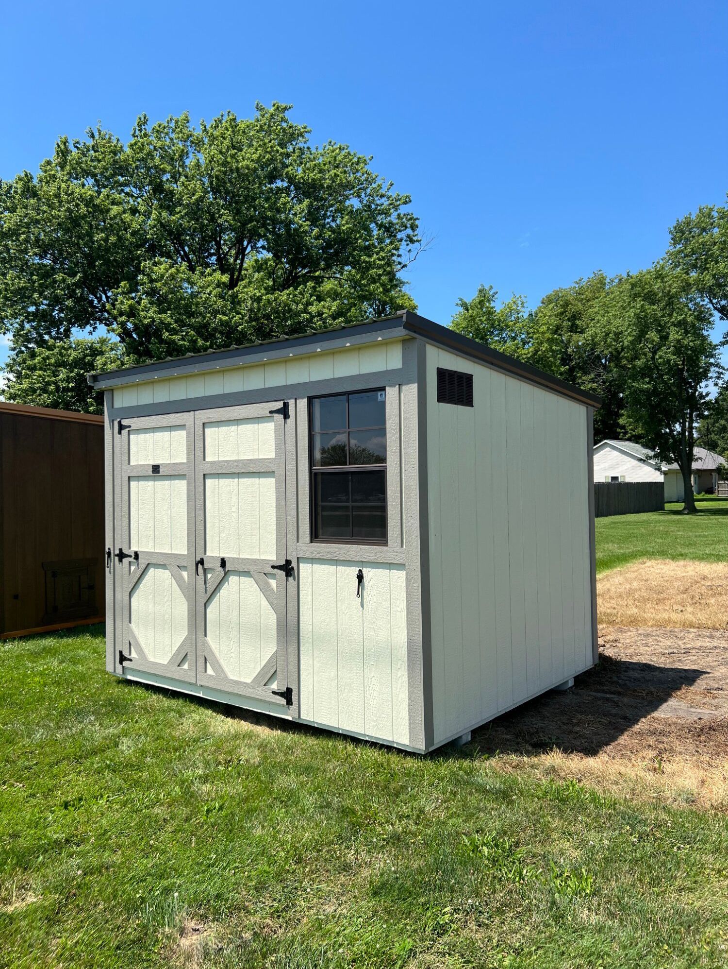 A light-colored shed with dark trim, a single window, and double doors, situated in a green grassy area.