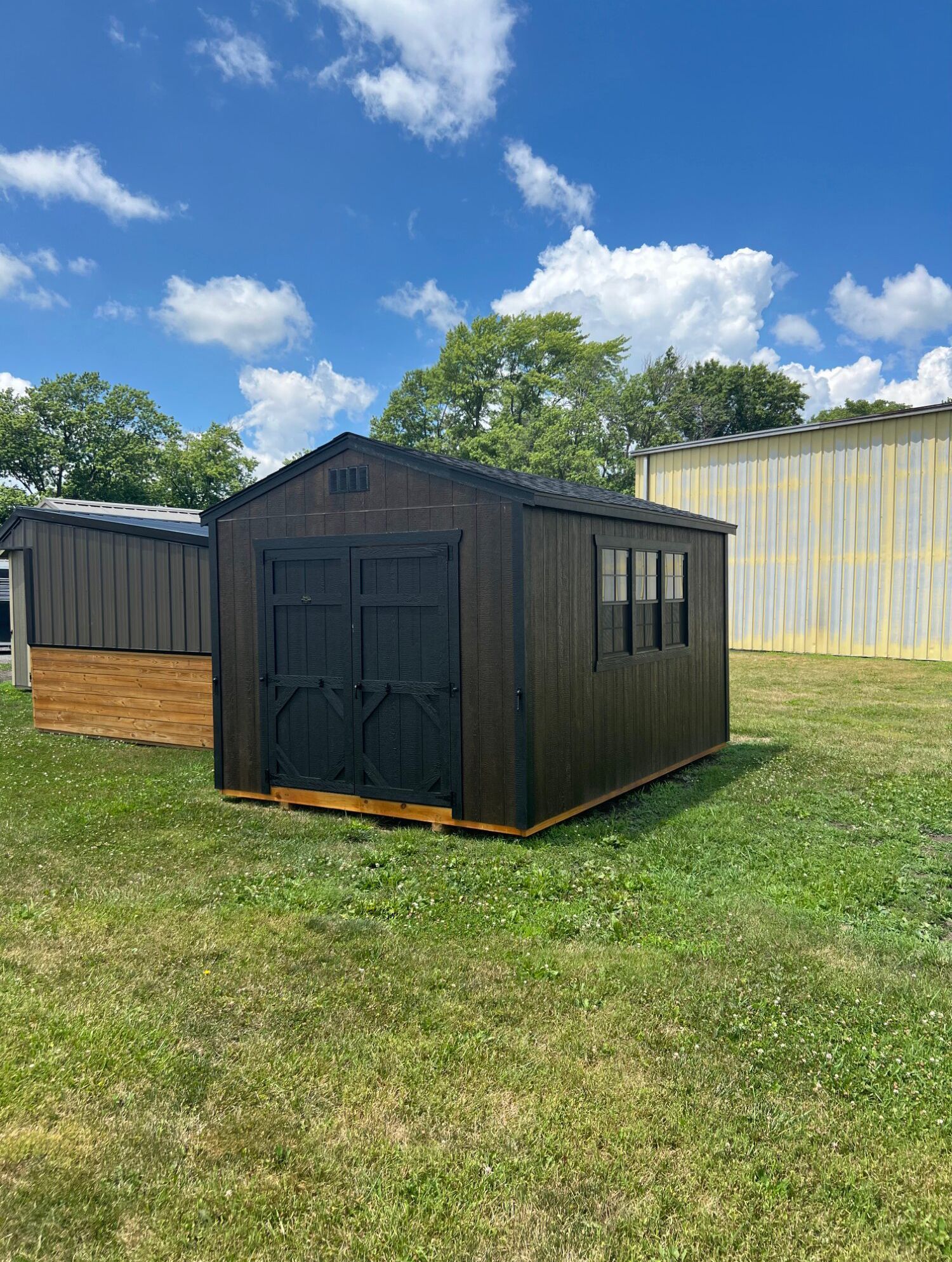 A dark brown wooden shed with black double doors and a window, sitting on a grassy lot under a bright blue sky.