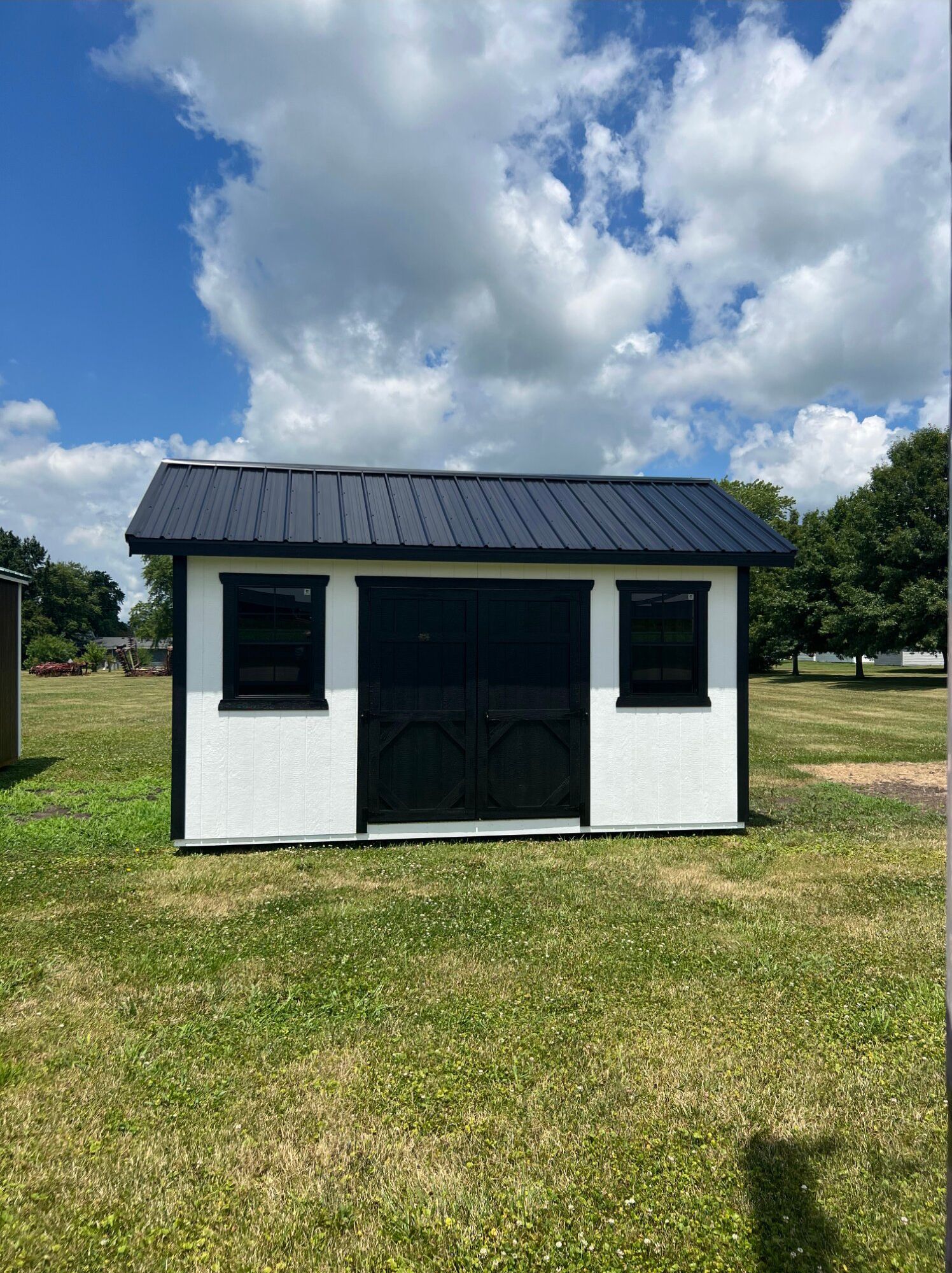 A white, rectangular shed with a black metal roof, black trim, black double doors, and two black windows in a grassy field.