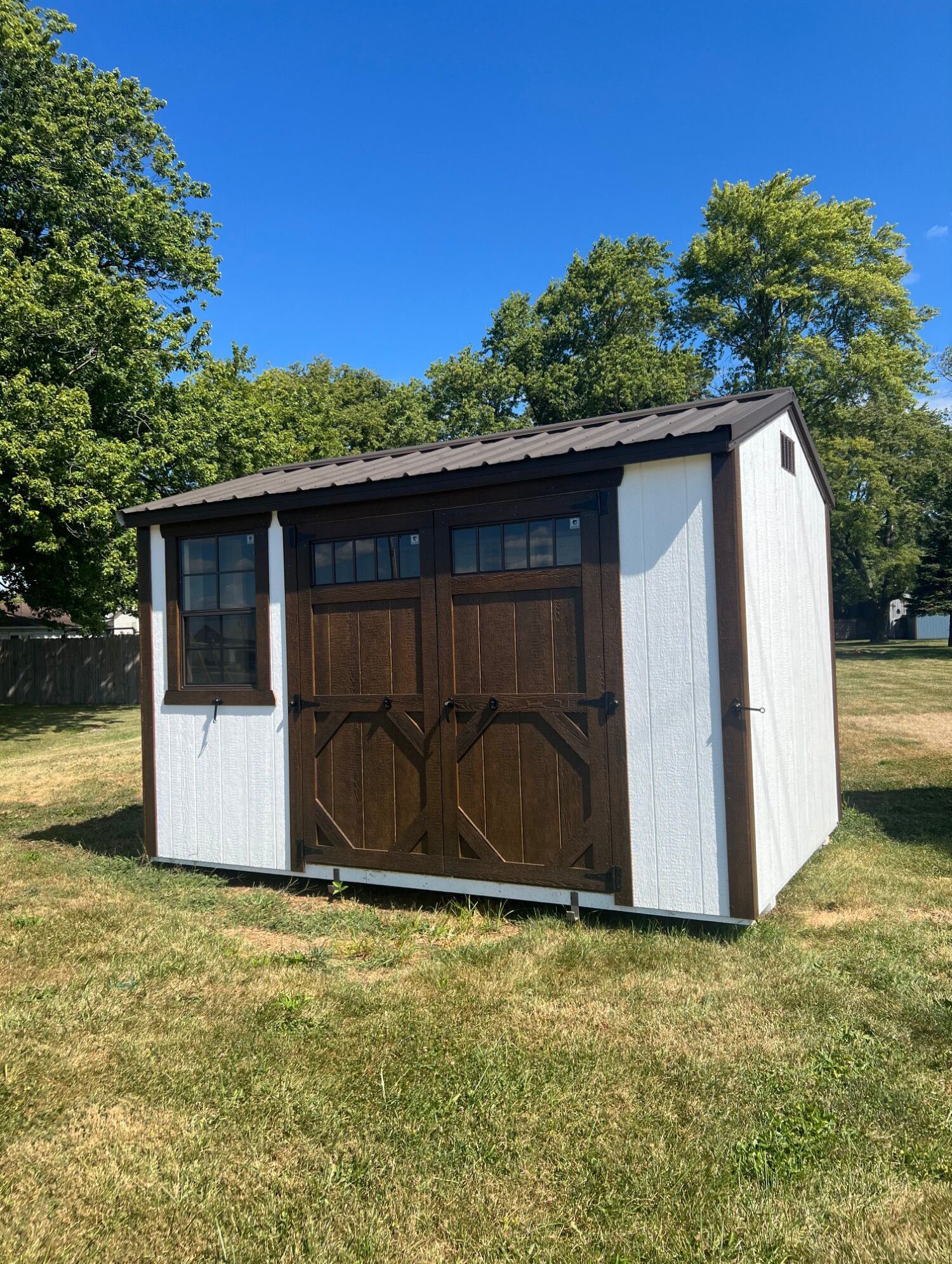 A white wooden shed with dark brown trim, double doors, and a side window, sitting in a grassy yard under a blue sky.
