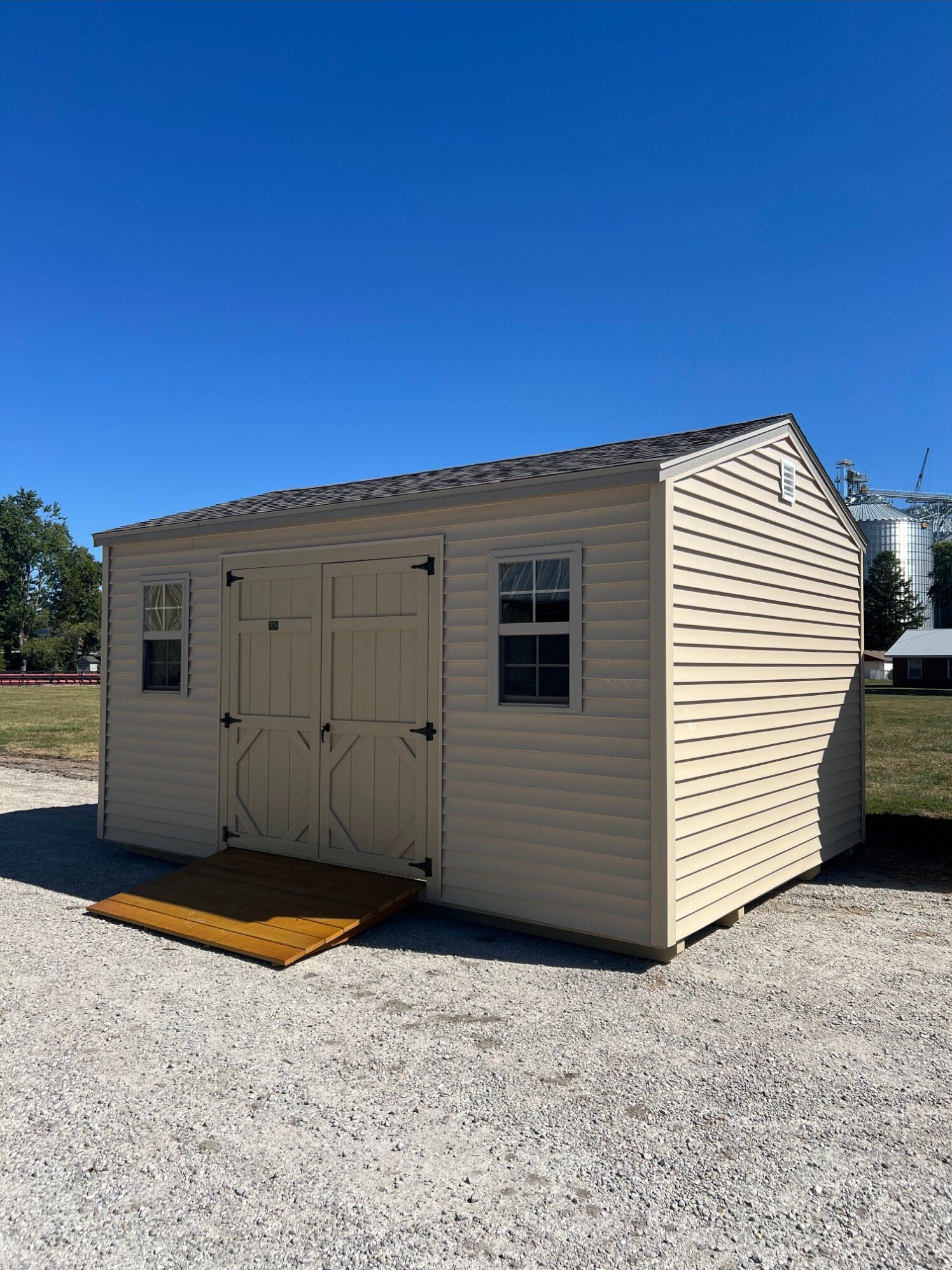 A tan, rectangular storage shed with double doors, two windows, and a ramp, sitting on a gravel lot under a clear sky.