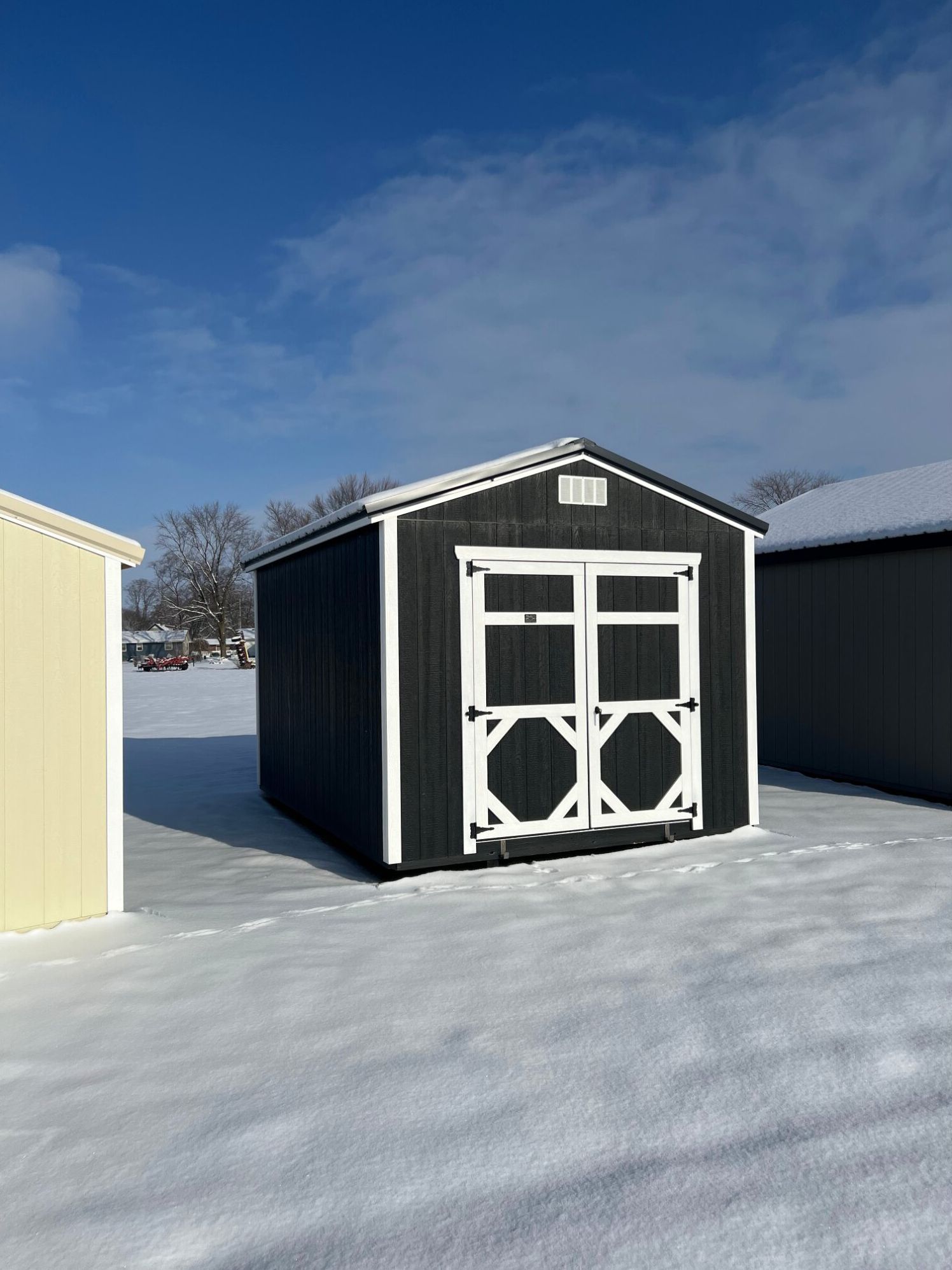 A dark gray wooden storage shed with white trim and double doors, standing in a snowy field under a bright blue sky.