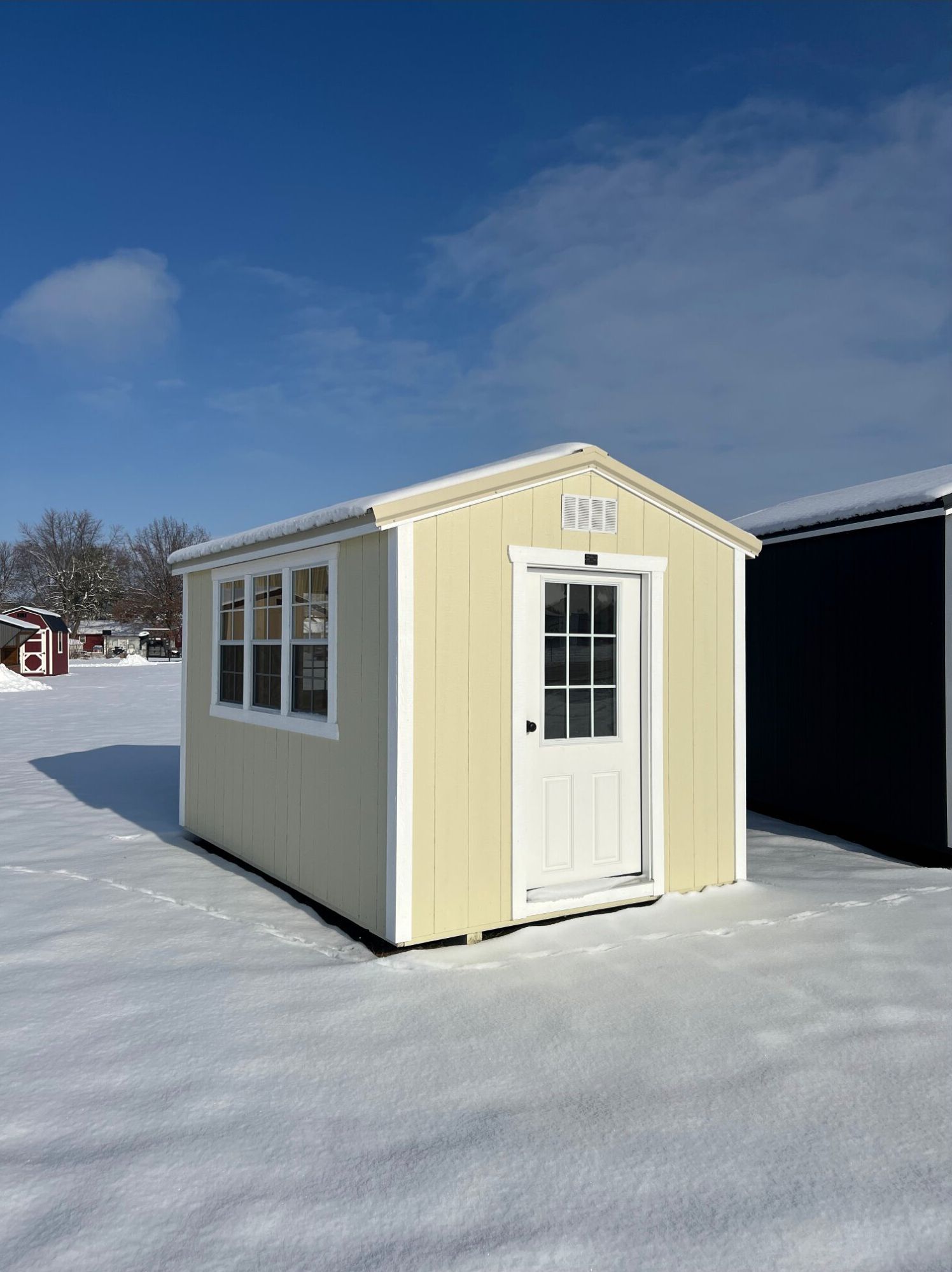A light yellow, small shed with white trim and a single windowed door, standing in a snowy field under a blue sky.