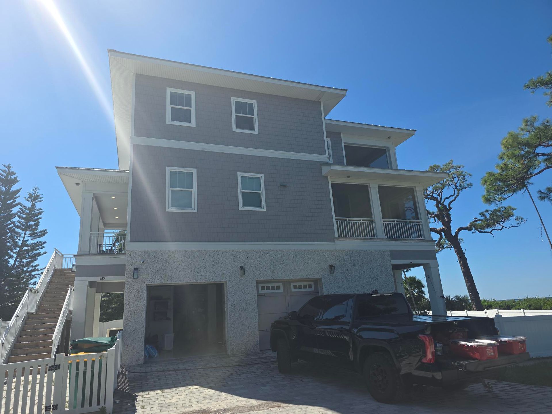 Three-story house with gray siding, white trim, and a truck parked in the driveway under a bright sky.