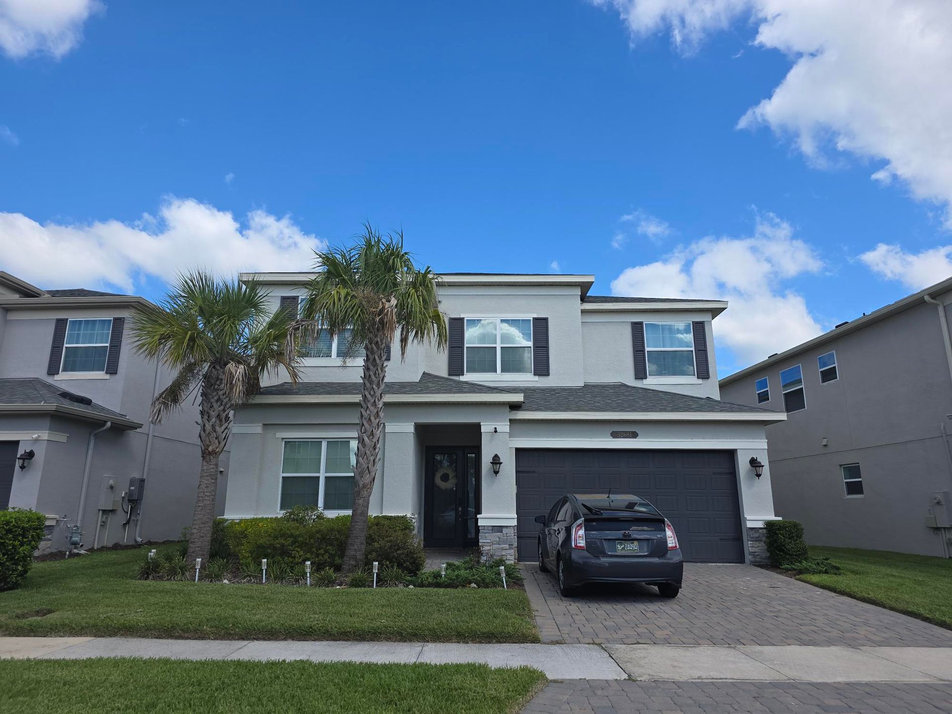 Two-story gray house with black shutters, palm trees, and a dark car in the driveway under a blue sky with clouds.