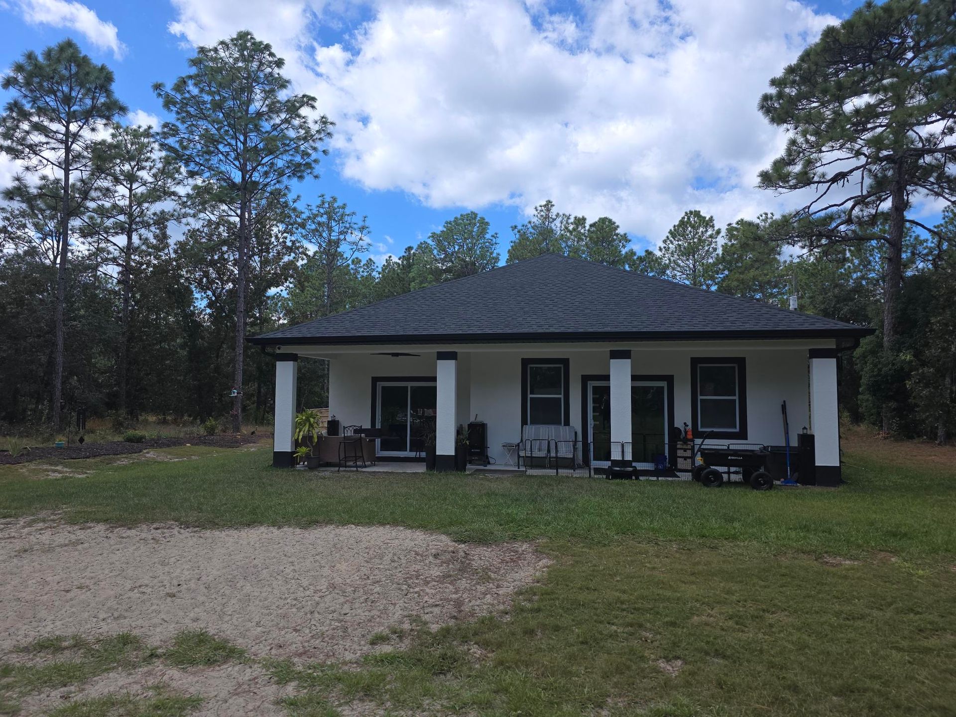 White house with black trim, porch supported by columns, surrounded by trees under a cloudy sky.