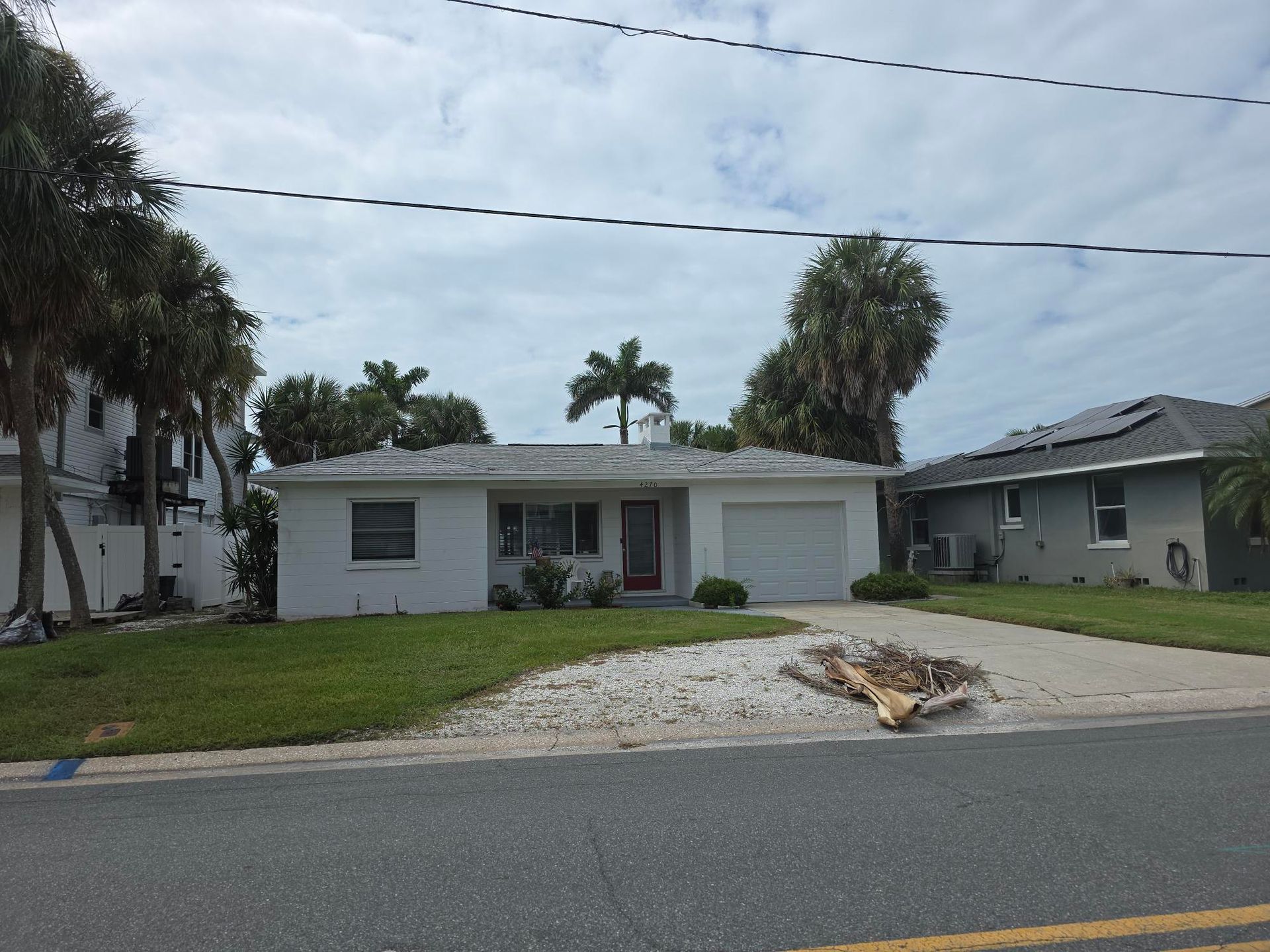 White house with attached garage, palm trees, and gravel driveway under cloudy sky.