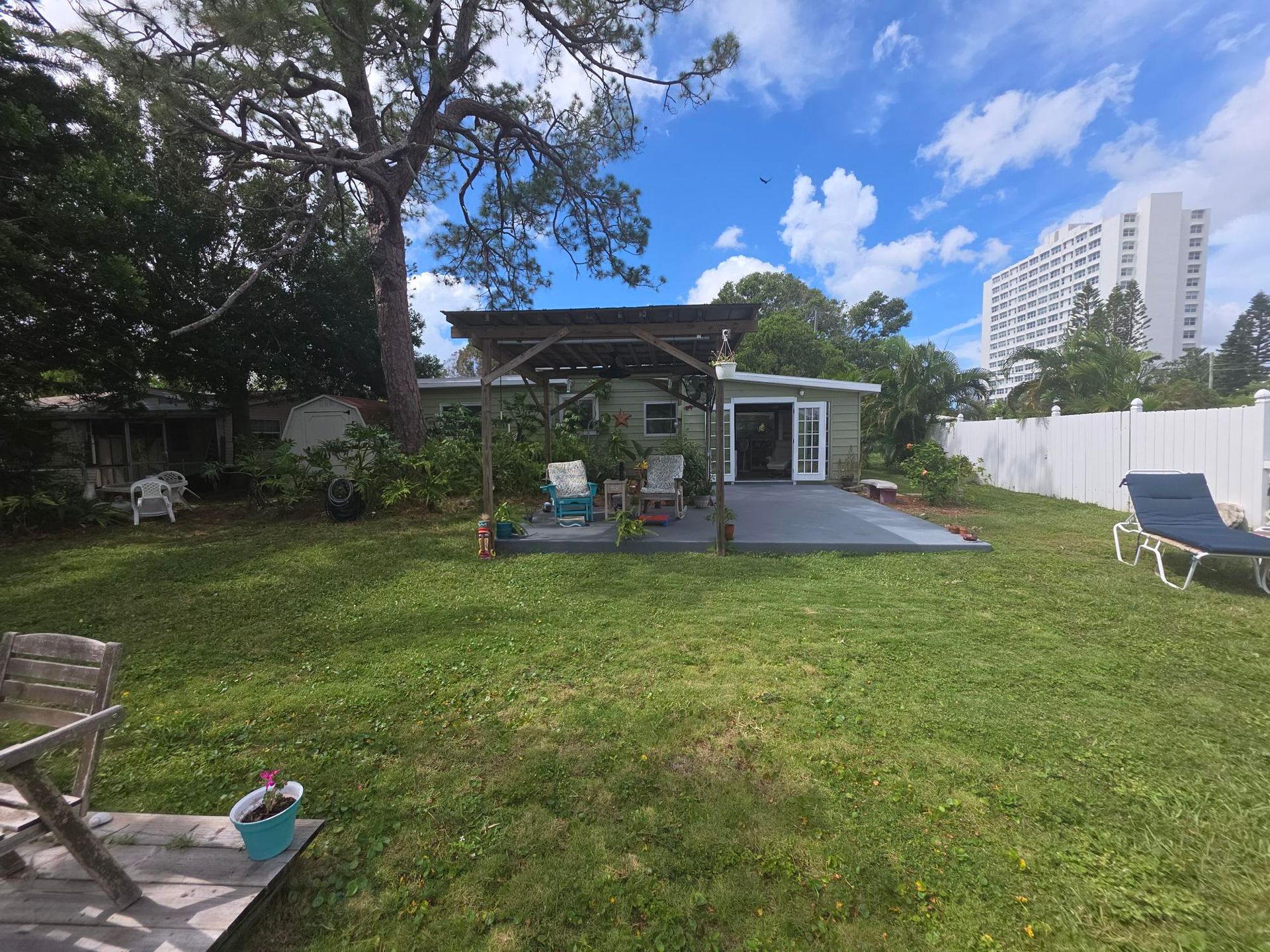Backyard with lawn, patio, small house, pergola, lounge chairs, and tall building in the distance.