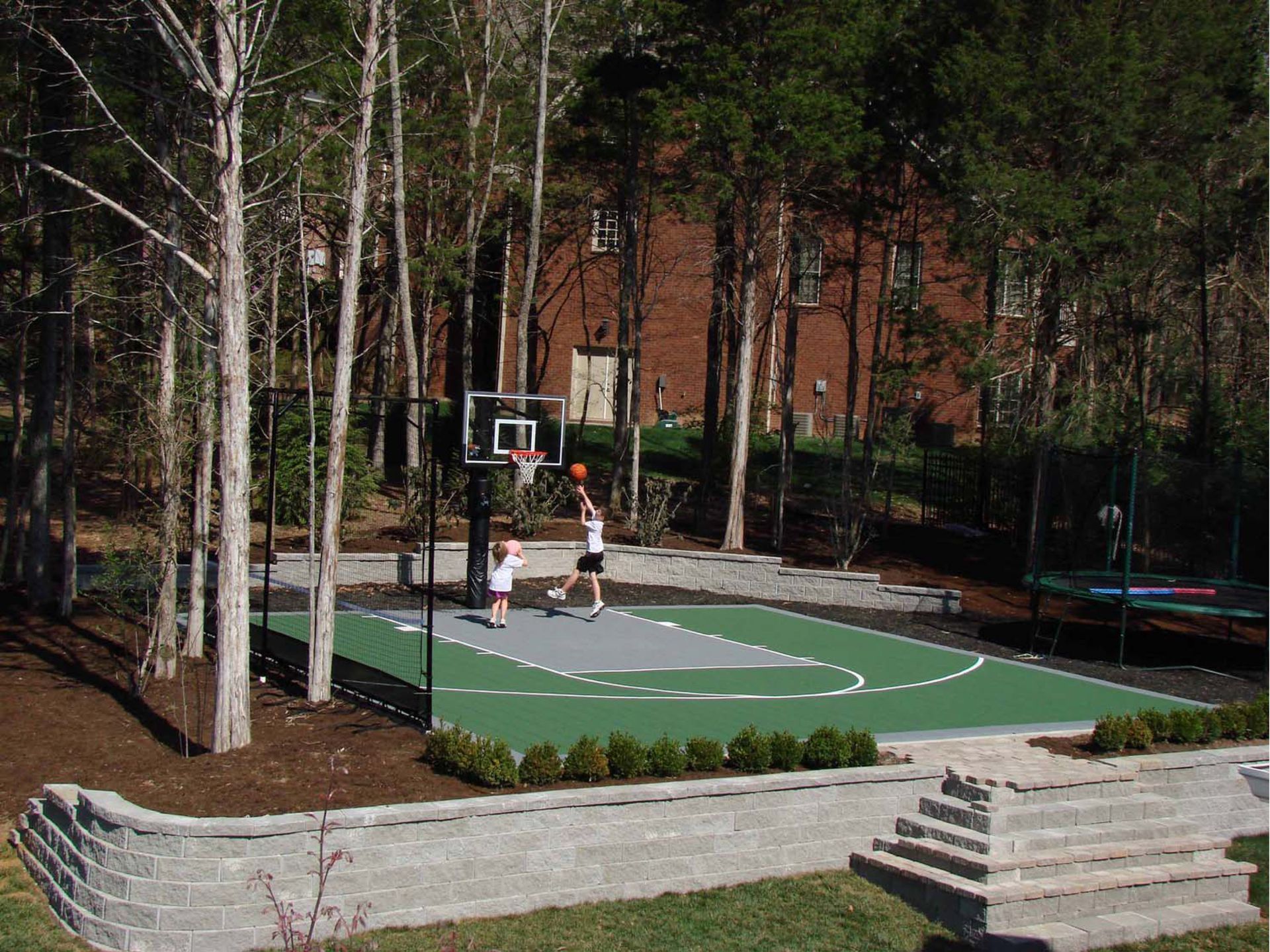 Two people are playing basketball in a backyard surrounded by trees