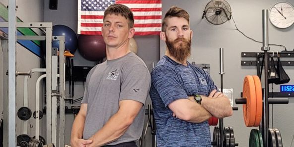 Two men standing back-to-back in a gym, arms crossed. American flag in the background.