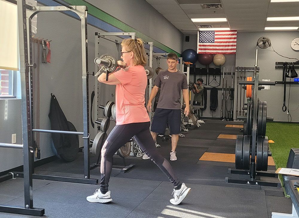 A woman is lifting a dumbbell in a gym while a man watches.
