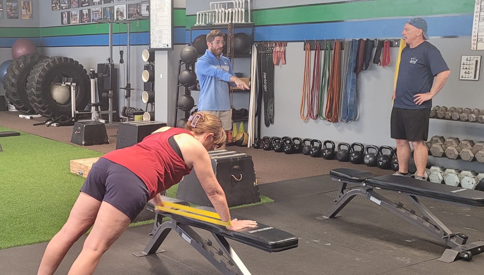 Woman stretching on bench in a gym while another person and a coach stand nearby.