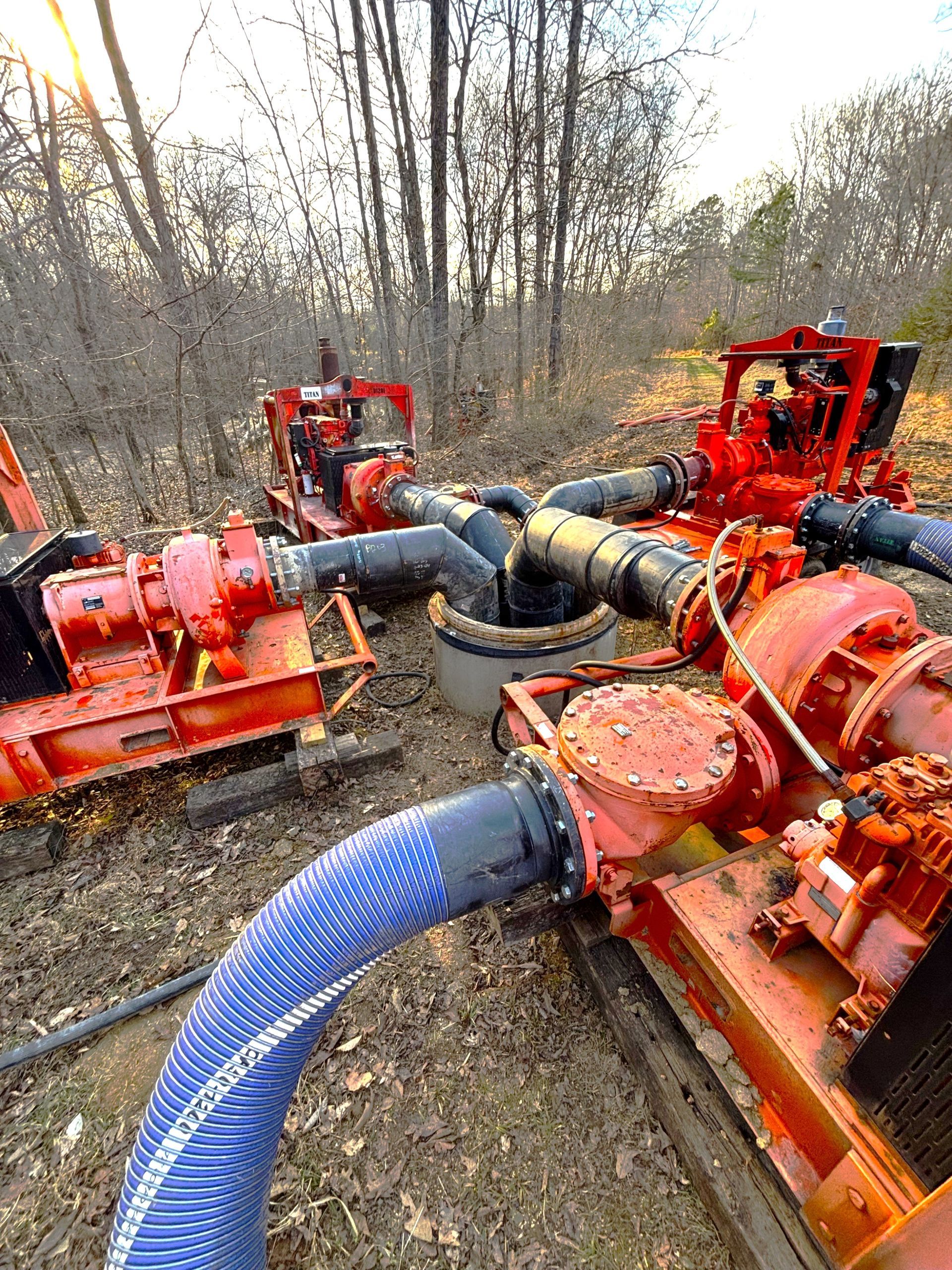 Several bright red industrial pumps connected by black pipes to a central hub in a wooded area.