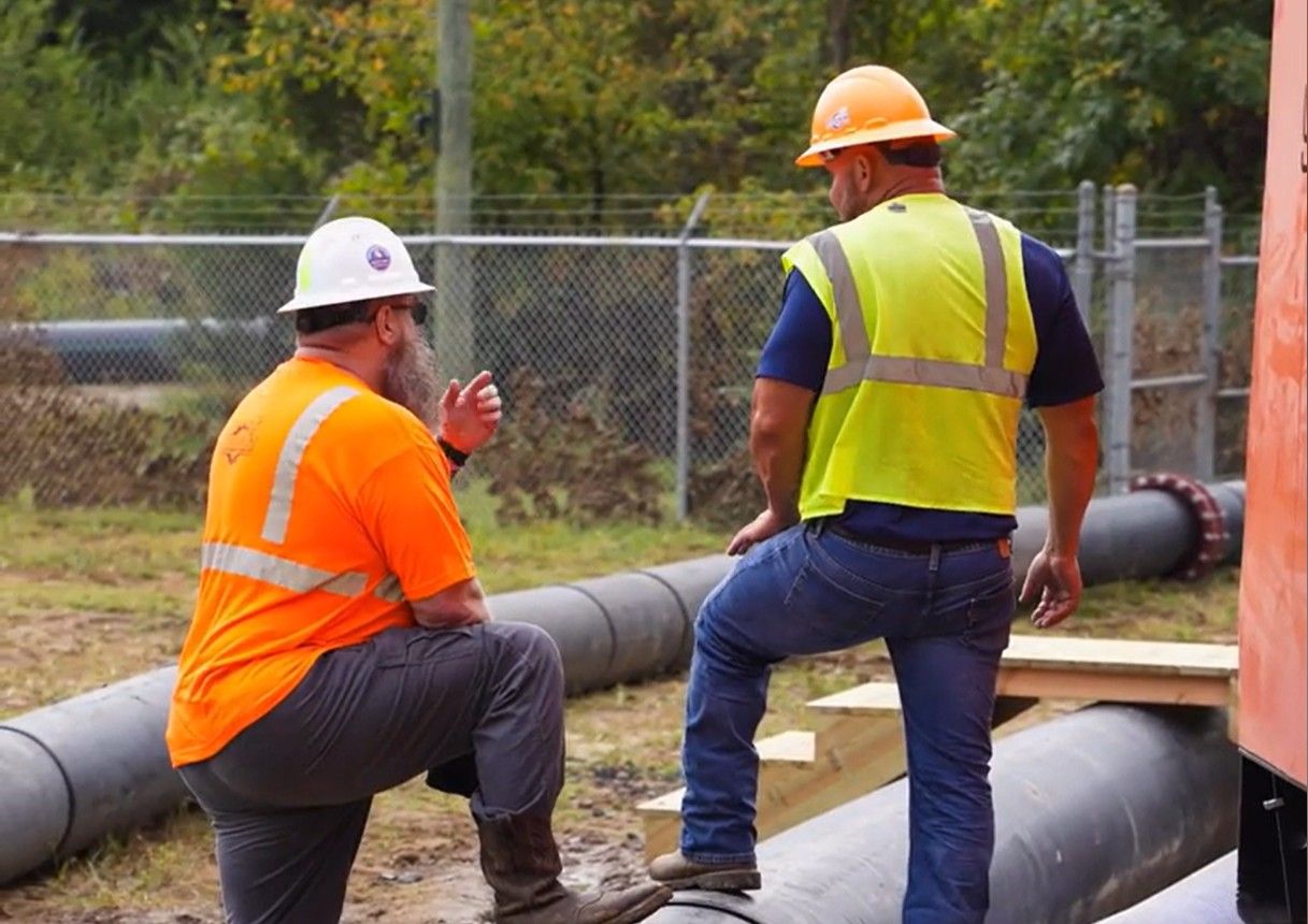 Two construction workers in safety gear and hard hats discuss work near large pipes at an outdoor site.