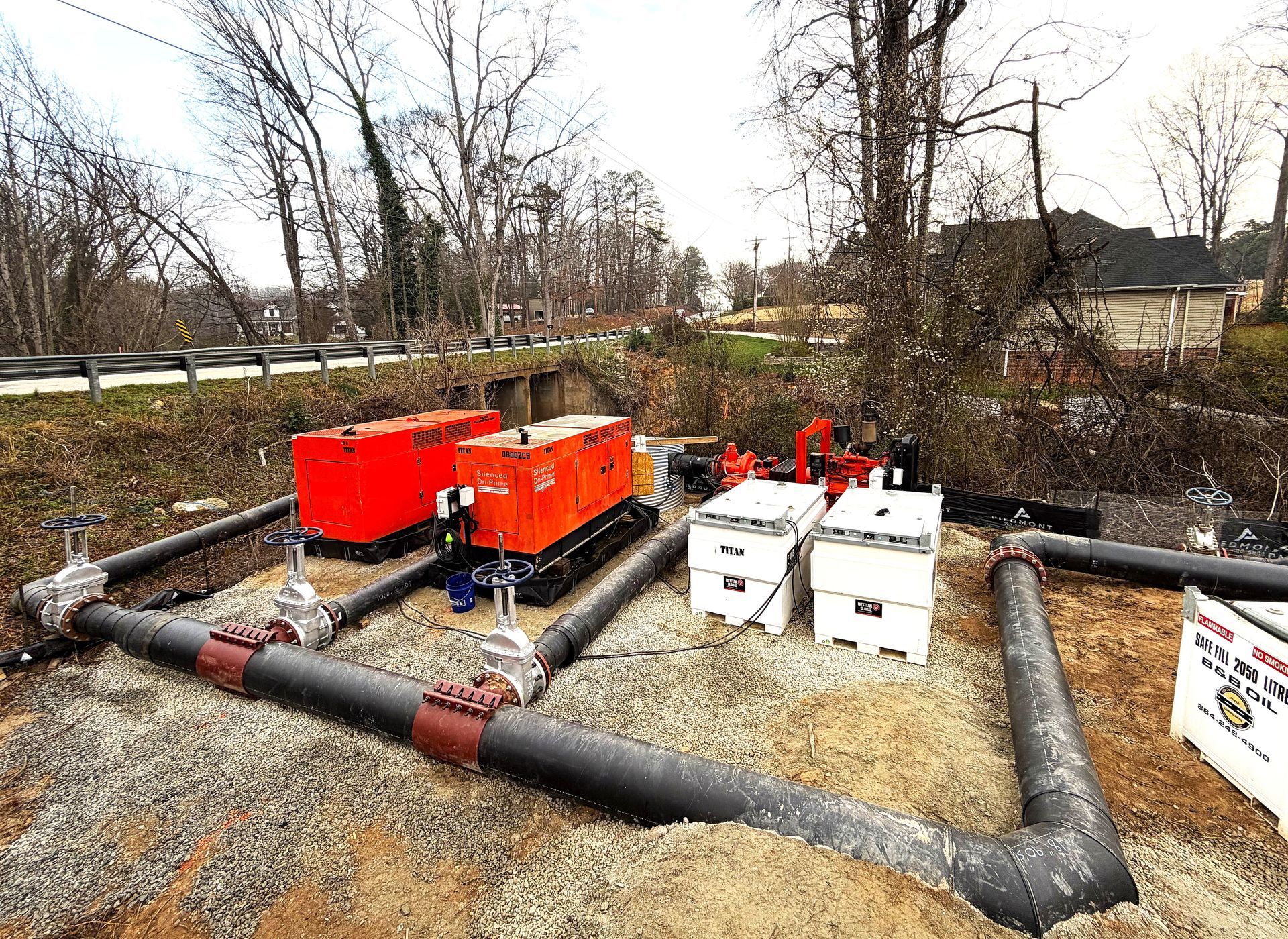 Two orange industrial pumps connected to black pipes on a gravel bed outdoors, with white fuel tanks nearby.