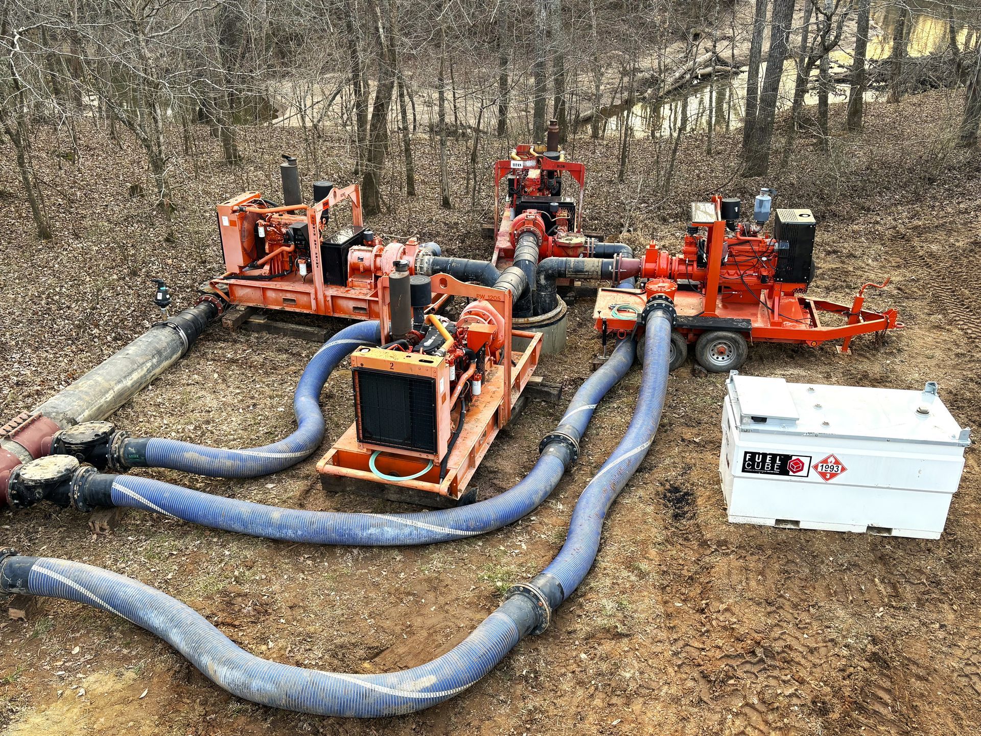 Three orange industrial pump units connected by blue hoses in a wooded area near a white fuel tank.
