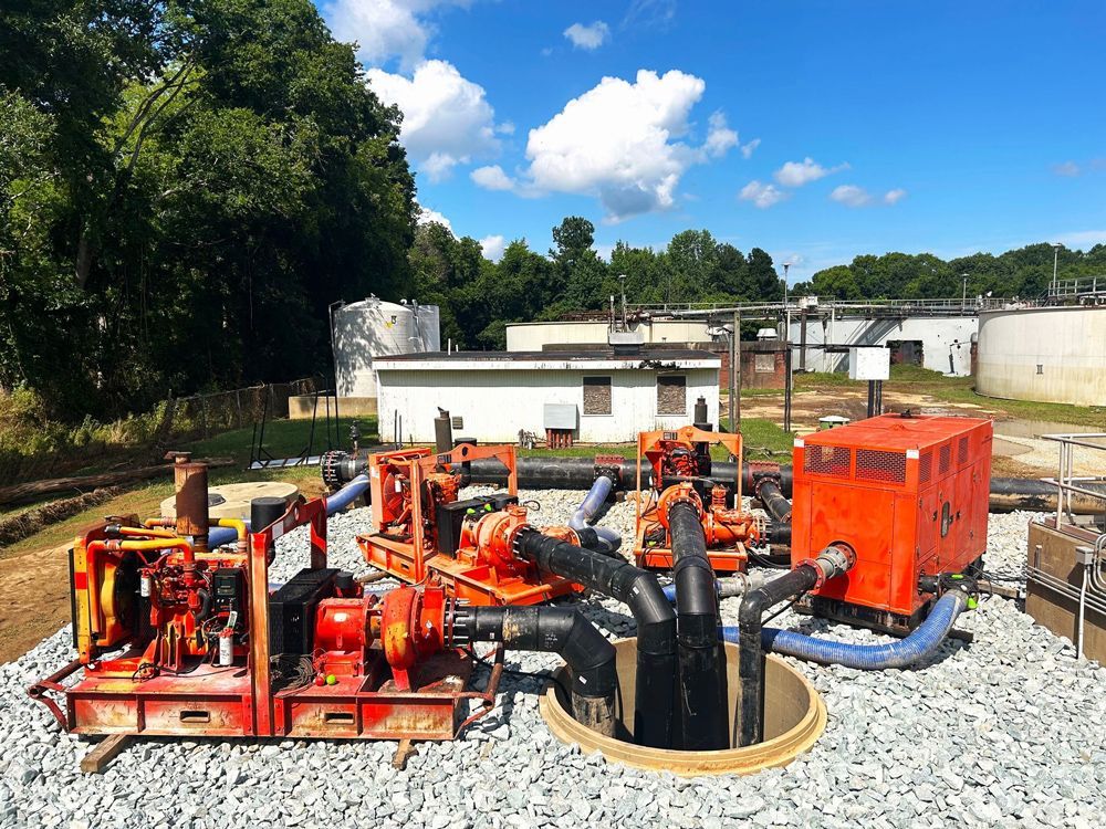 Orange industrial pumps and equipment in a gravel lot with tanks and buildings in the background under a blue sky.