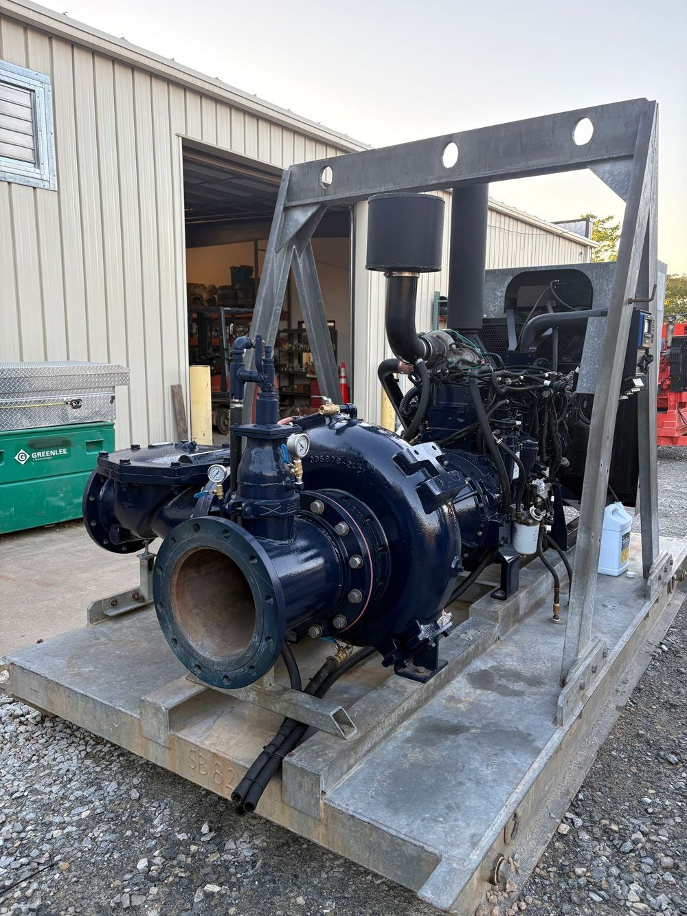 A large, dark blue water pump with an engine is mounted on a wooden pallet outside a building. It sits within a gray metal frame.