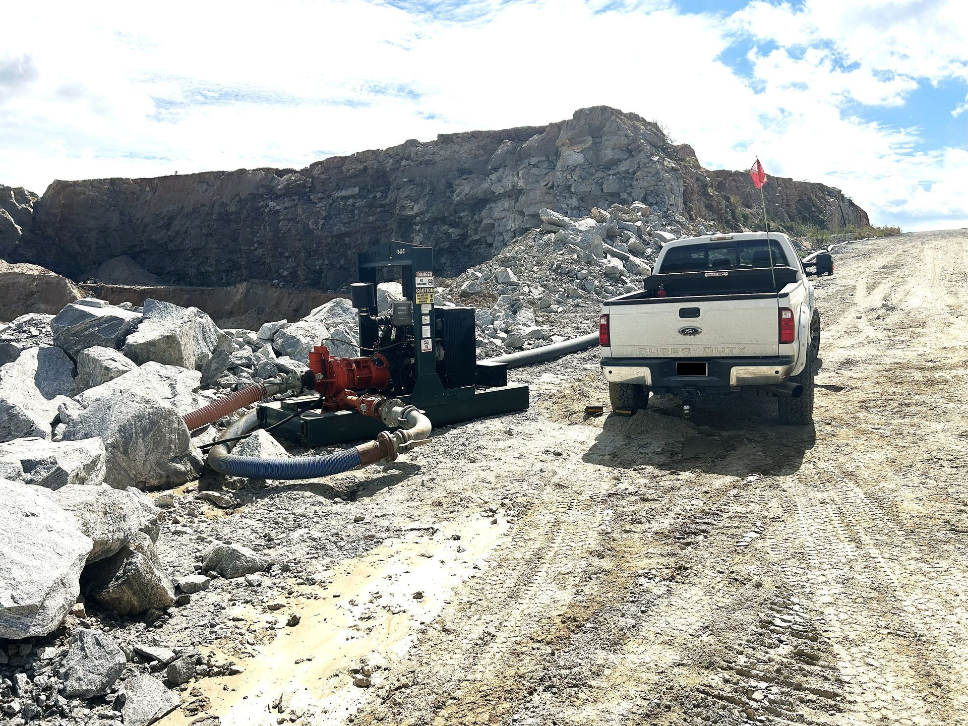A drilling machine and a white truck on a rocky terrain with a large rock formation in the background.