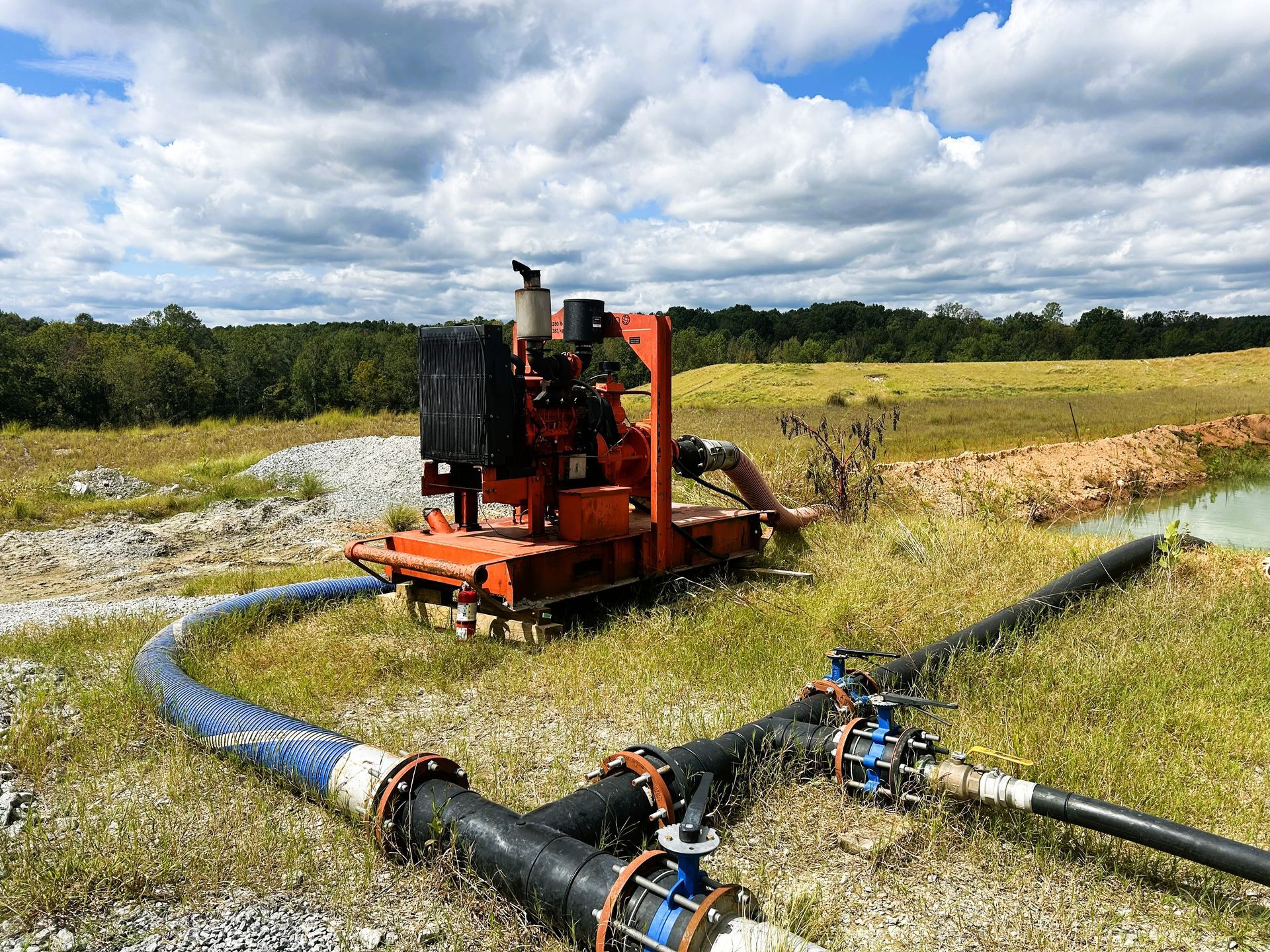 Orange water pump in a grassy field with blue and black pipes, under a cloudy sky.