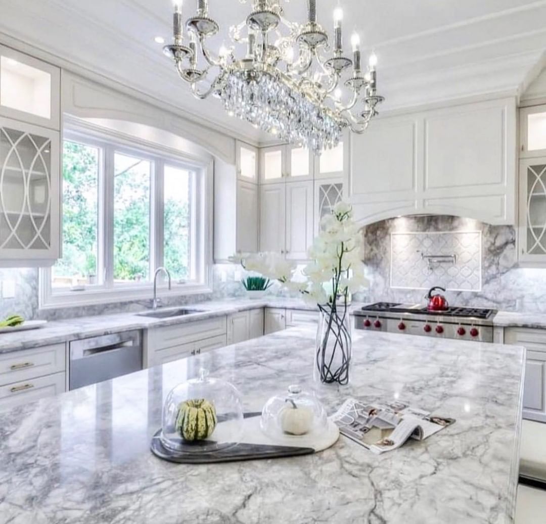 Elegant white kitchen with marble countertops, chandelier, and stainless steel appliances.