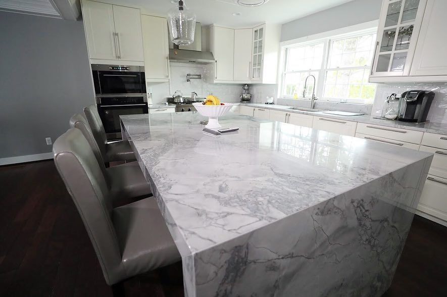 Modern kitchen with marble island, gray chairs, white cabinets, and stainless steel appliances.