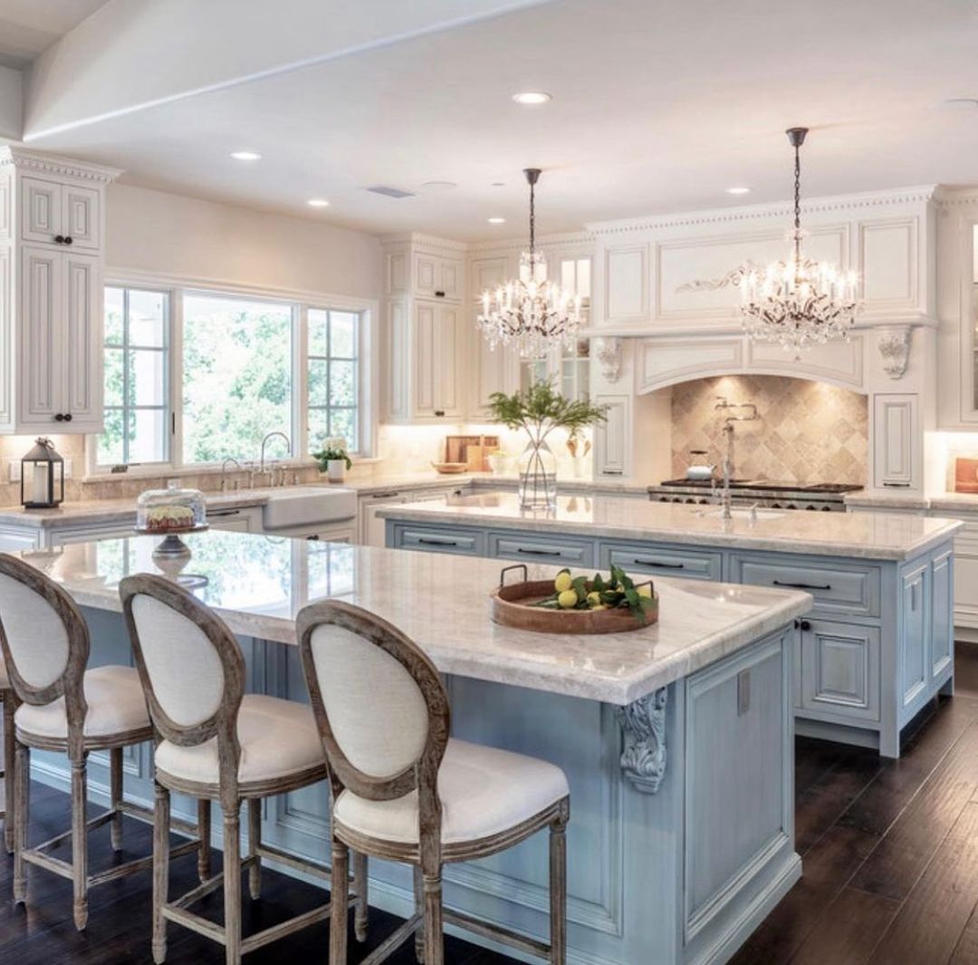 Elegant kitchen with light blue island, marble countertops, chandeliers, and bar stools.