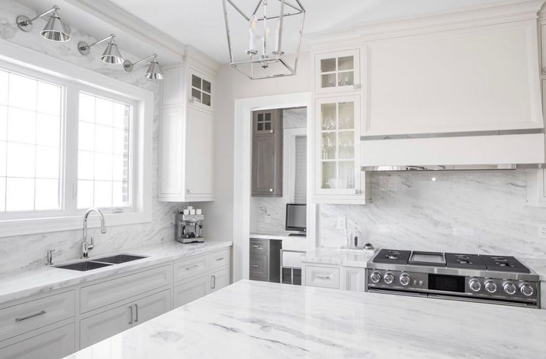 Modern white kitchen with marble countertops, stainless steel appliances, and a large window.