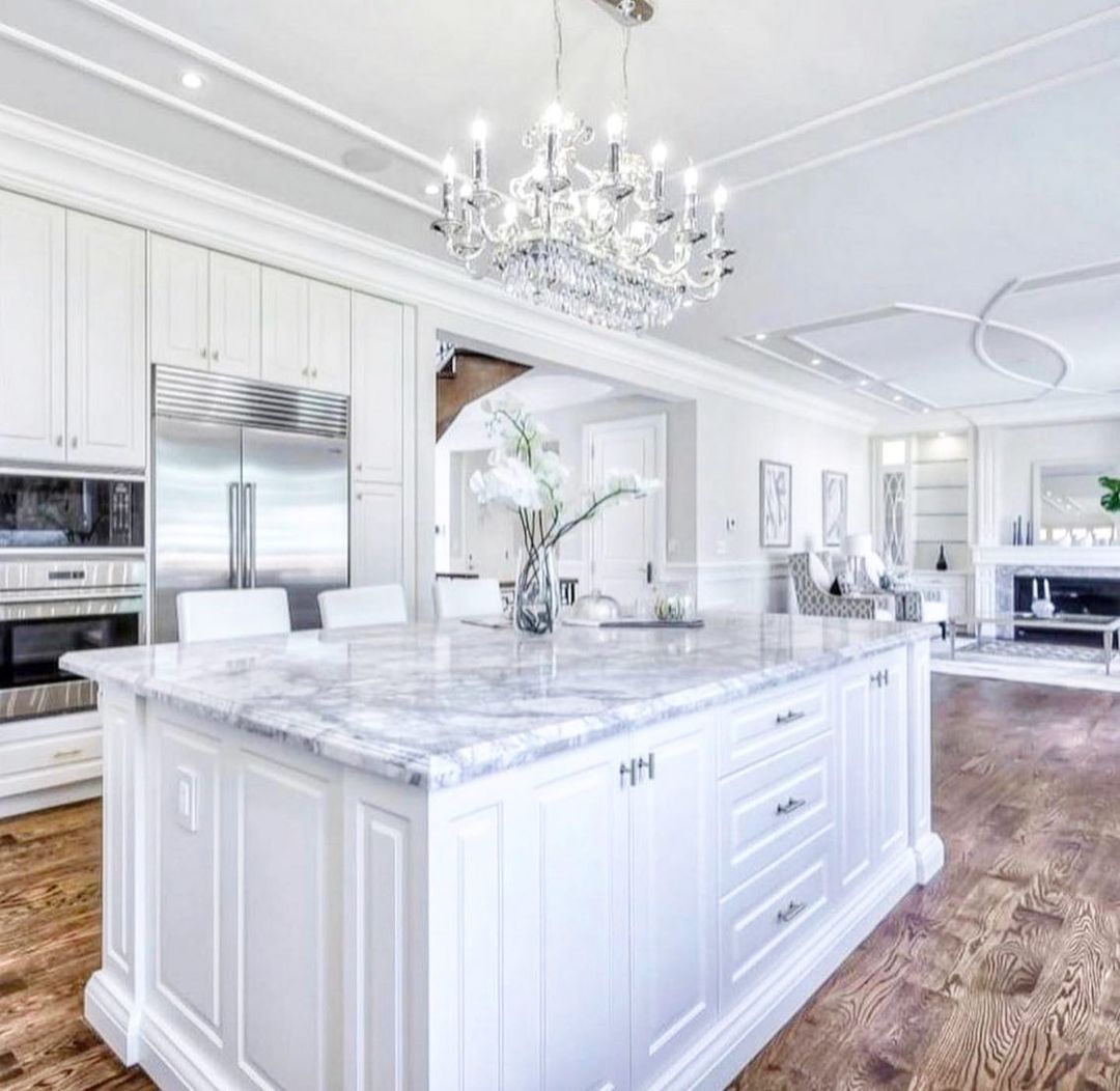 Luxurious white kitchen with marble island, chandelier, stainless steel appliances, and hardwood floors.