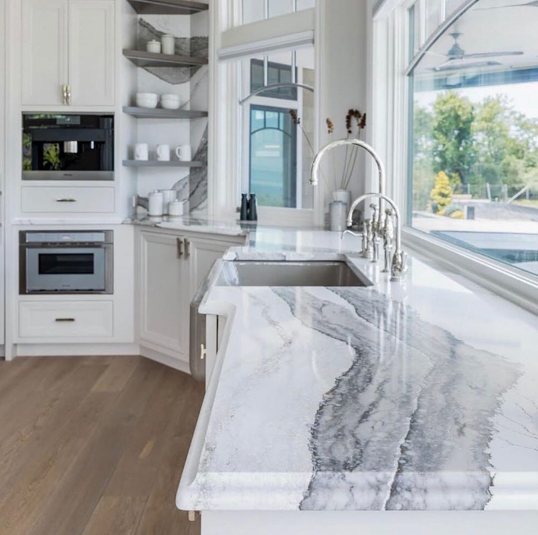 White kitchen with marble countertops, stainless steel sink, built-in appliances, and large windows with a view.