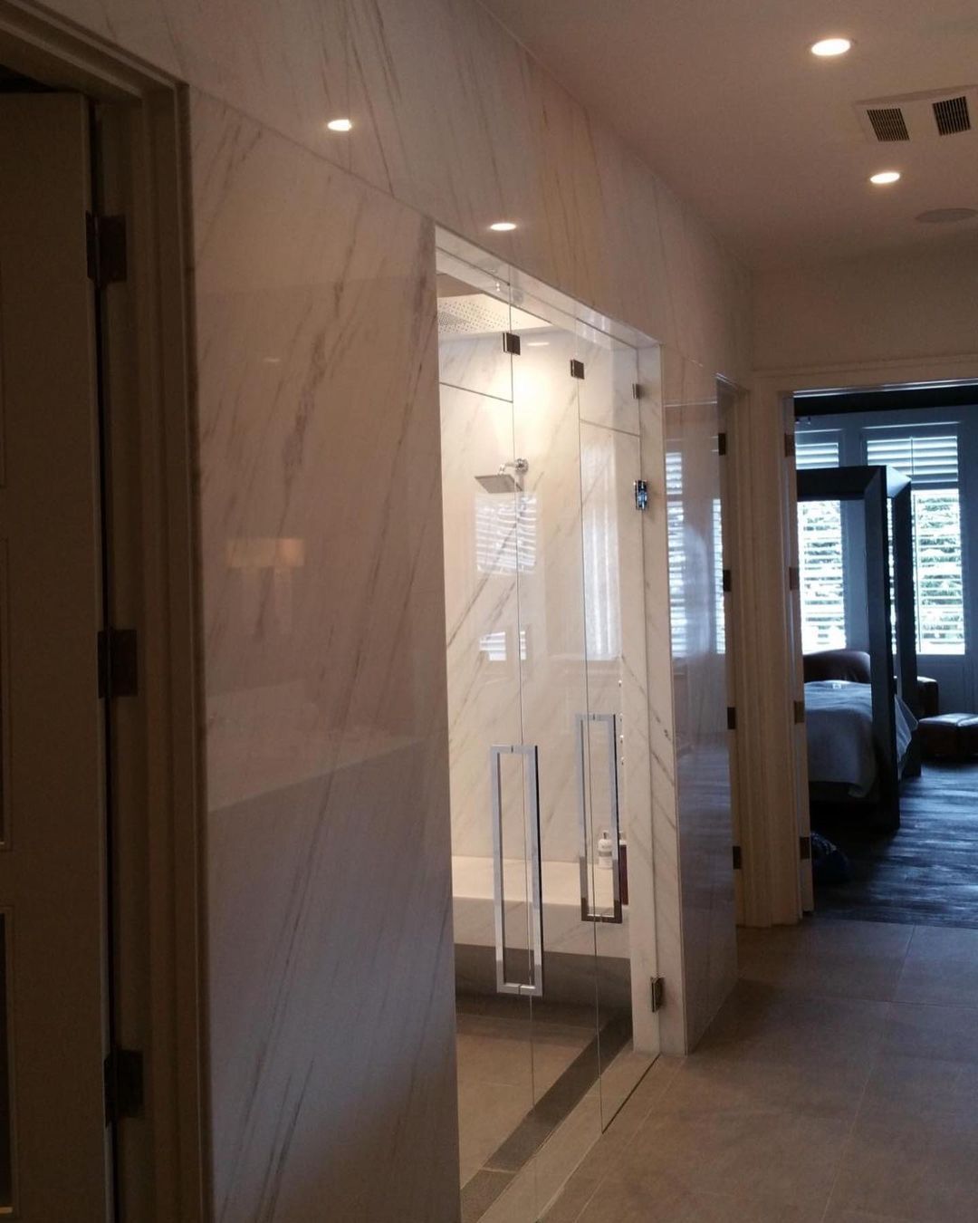 Luxurious marble-tiled bathroom with a glass shower and an open doorway leading to a bedroom.