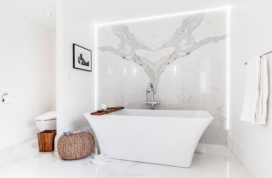 White bathroom with a freestanding tub against a marble accent wall, framed by a soft light.