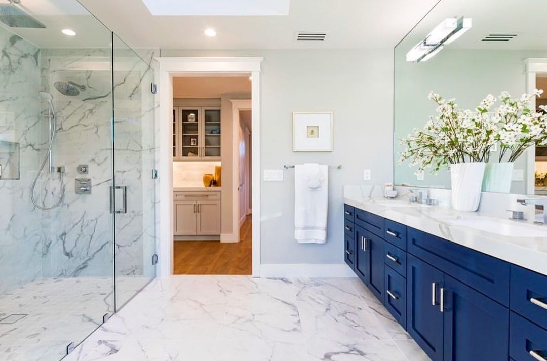 Modern bathroom with blue vanity, marble tile, glass shower, and open doorway to another room.