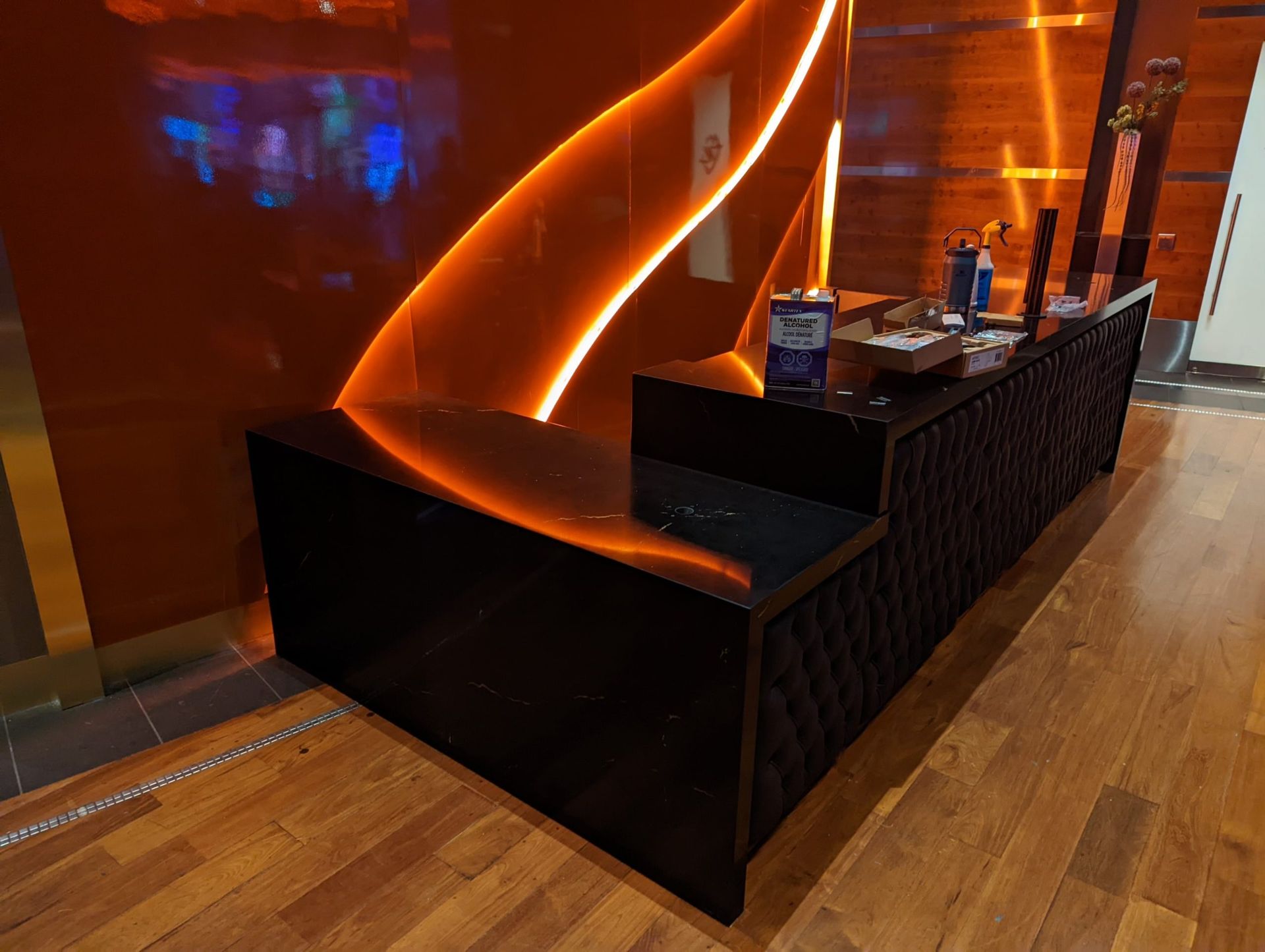 Black and shiny reception desk with diamond-patterned paneling, wooden floor, orange wall with glowing curve.
