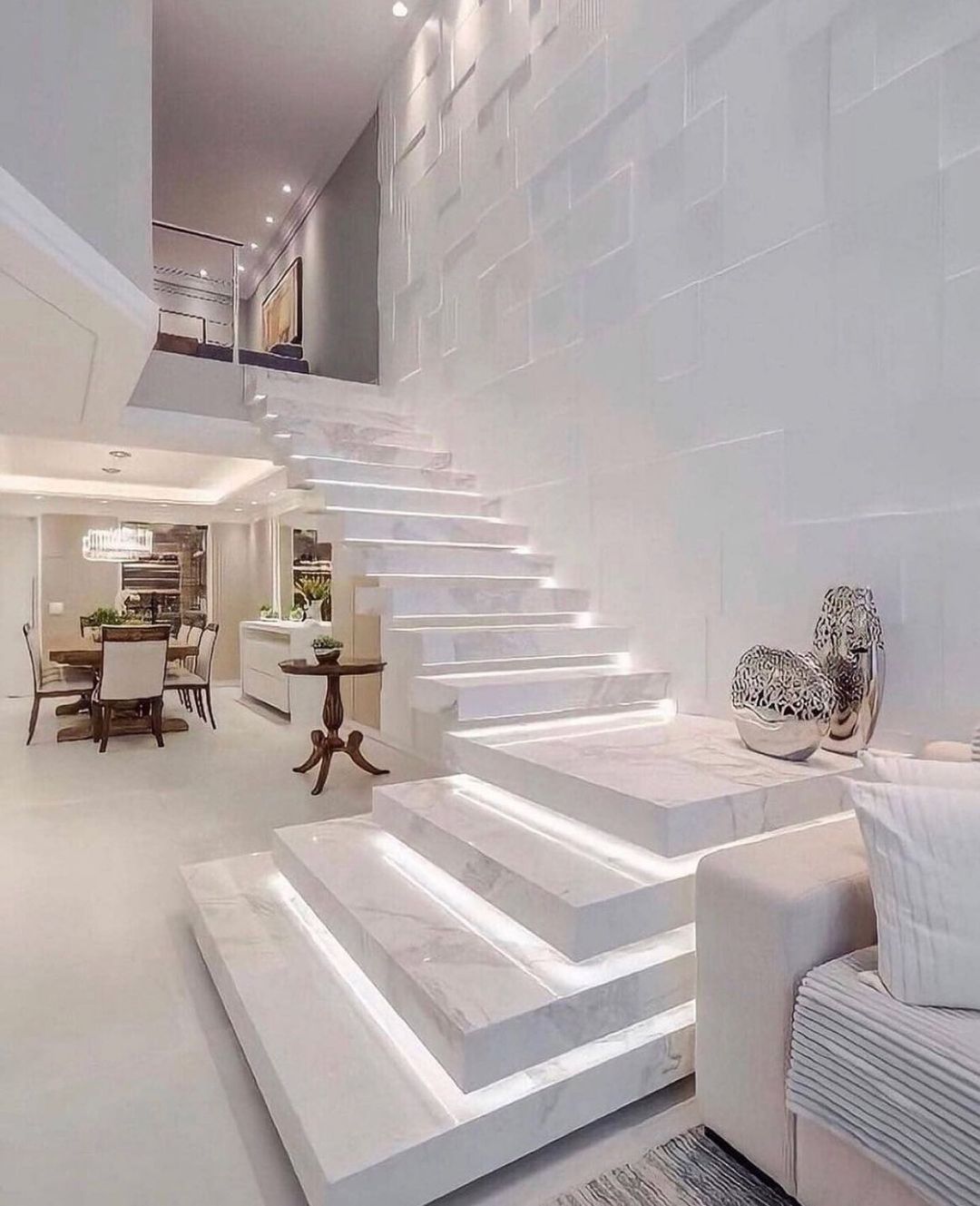 Spacious white marble staircase with under-step lighting, leading to a second floor. A living and dining area is visible.