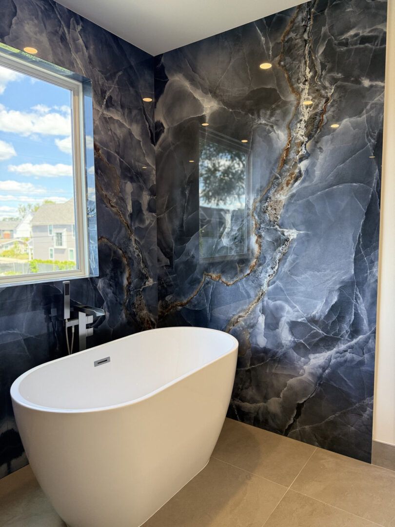 Modern bathroom with freestanding white tub, black marble walls, beige floor, and a window with sky view